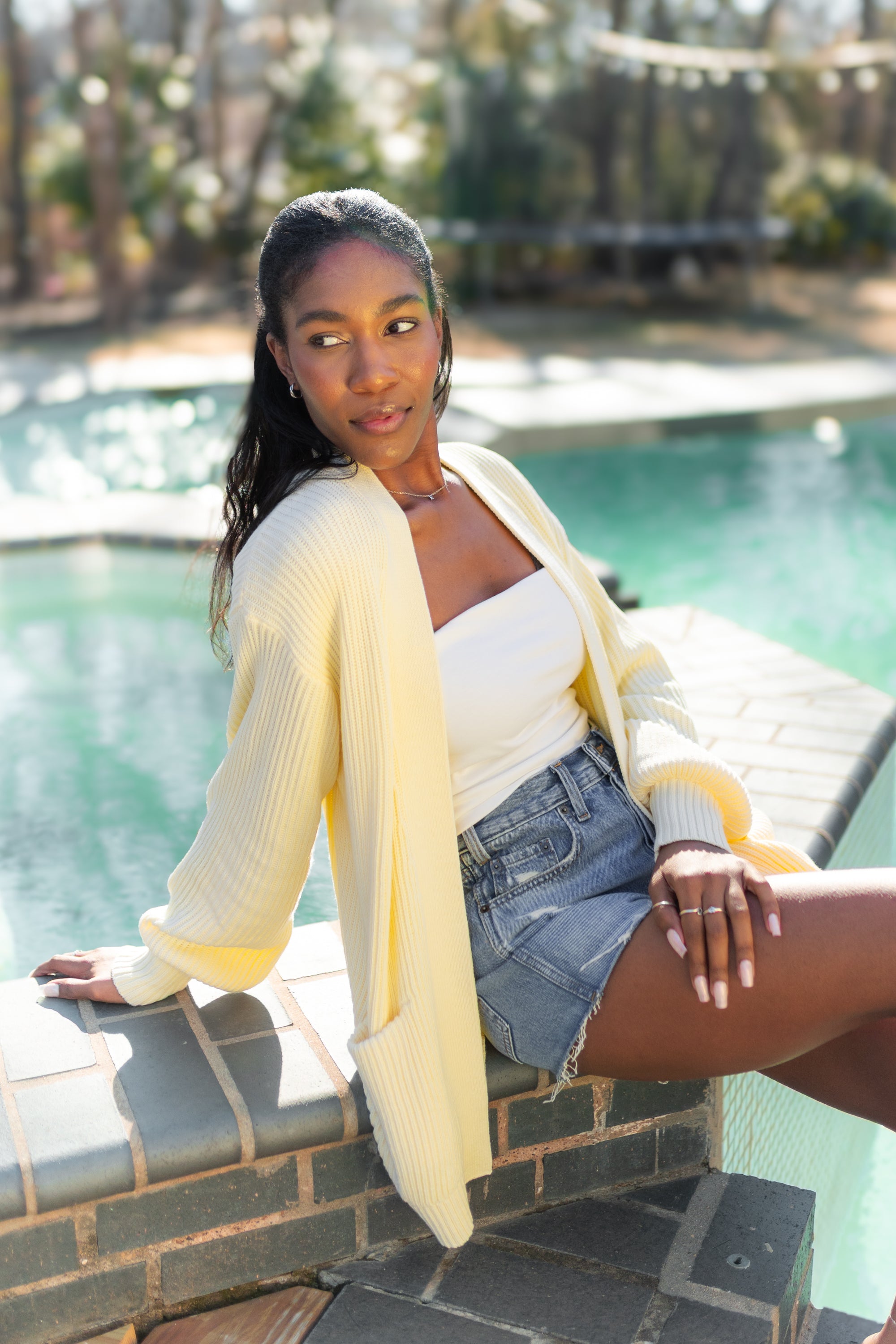 Female model sitting pool side wearing the Chunky Knit Women's Oversized Cardigan in Chamomile overtop a white shirt and medium wash jean shorts