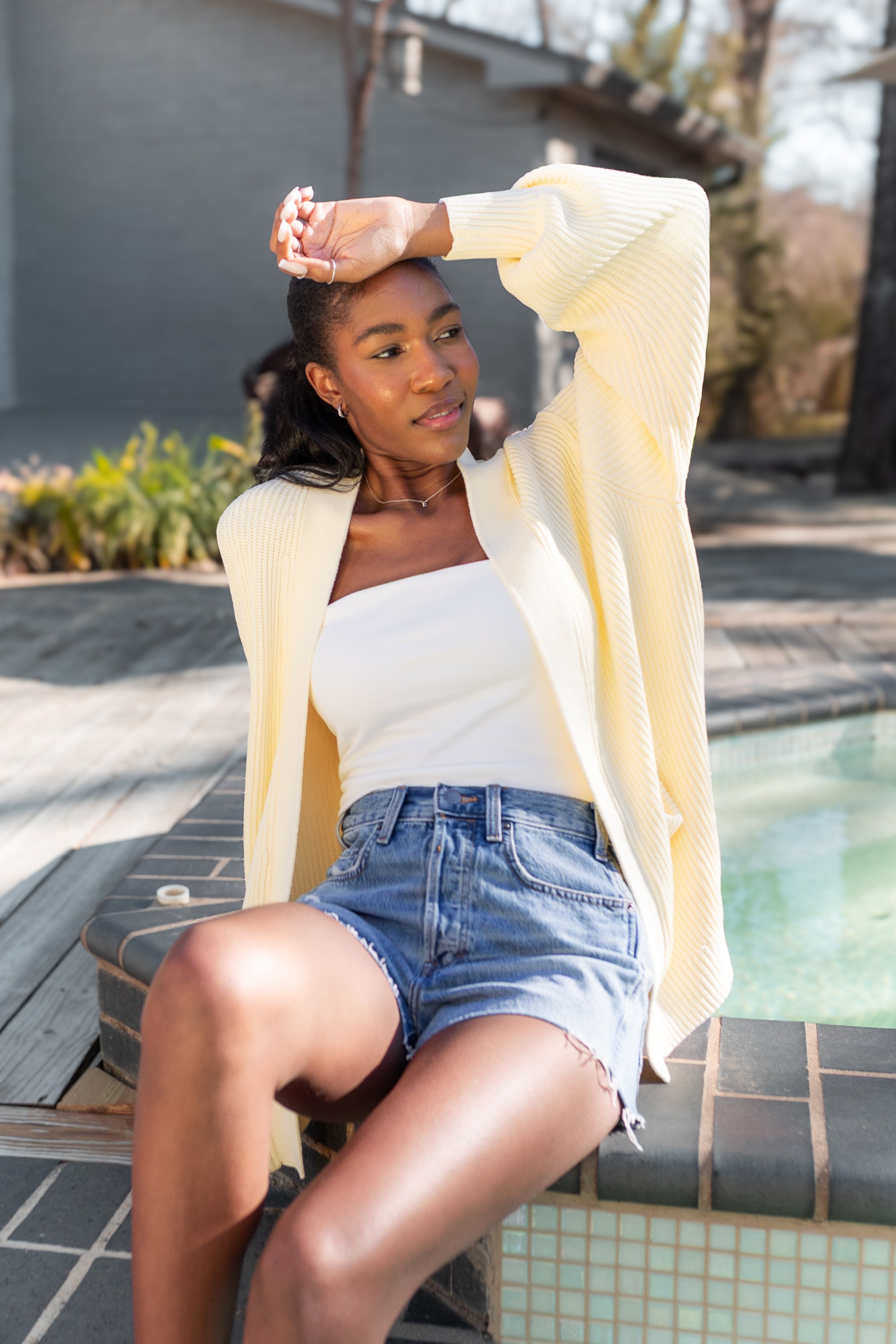 Female model sitting pool side with her hand on her forehead wearing the Chunky Knit Women's Oversized Cardigan in Chamomile with a white shirt and medium wash jean shorts
