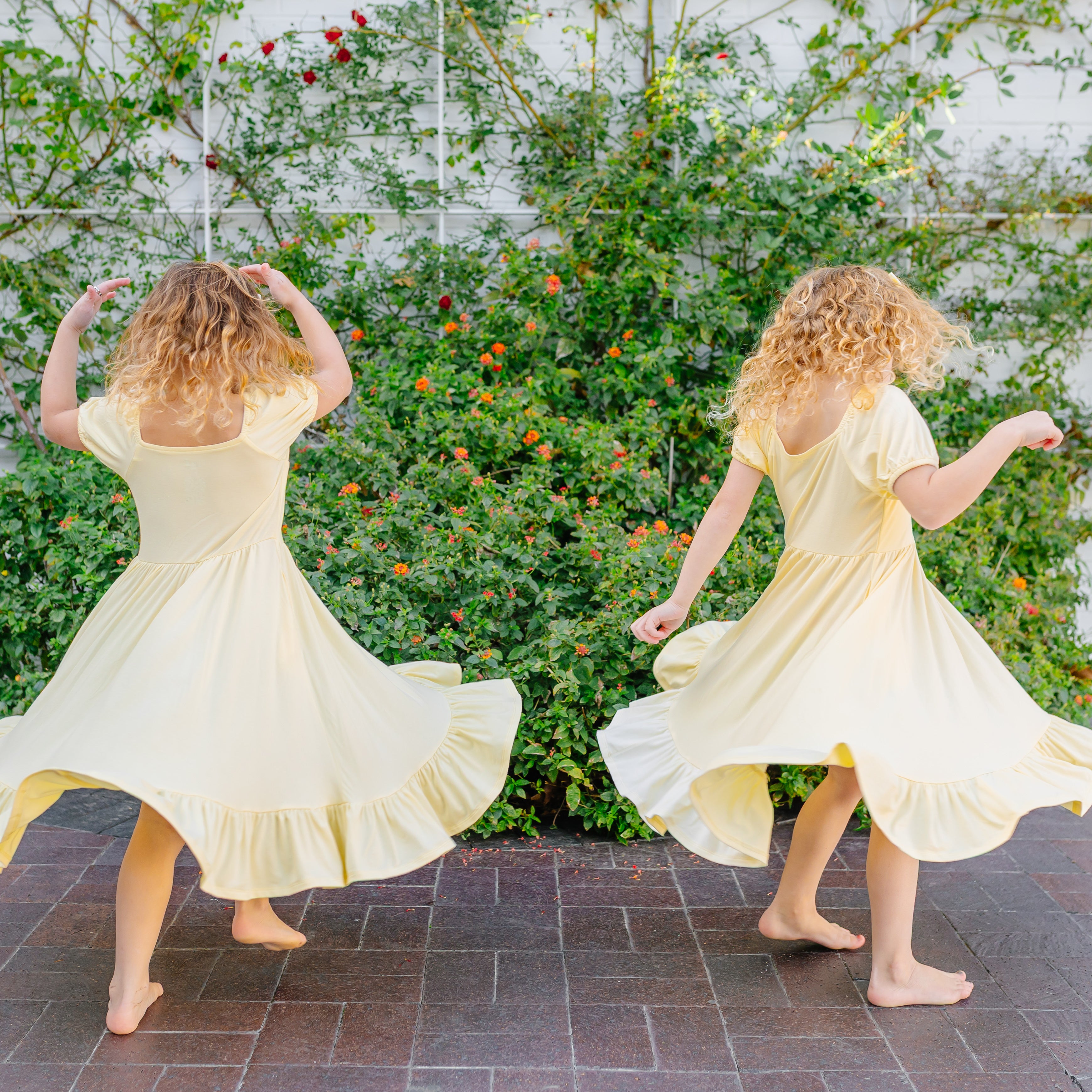 Two Girls wearing the Puff Sleeve Twirl Dress in Chamomile twirling outside in front of greenery