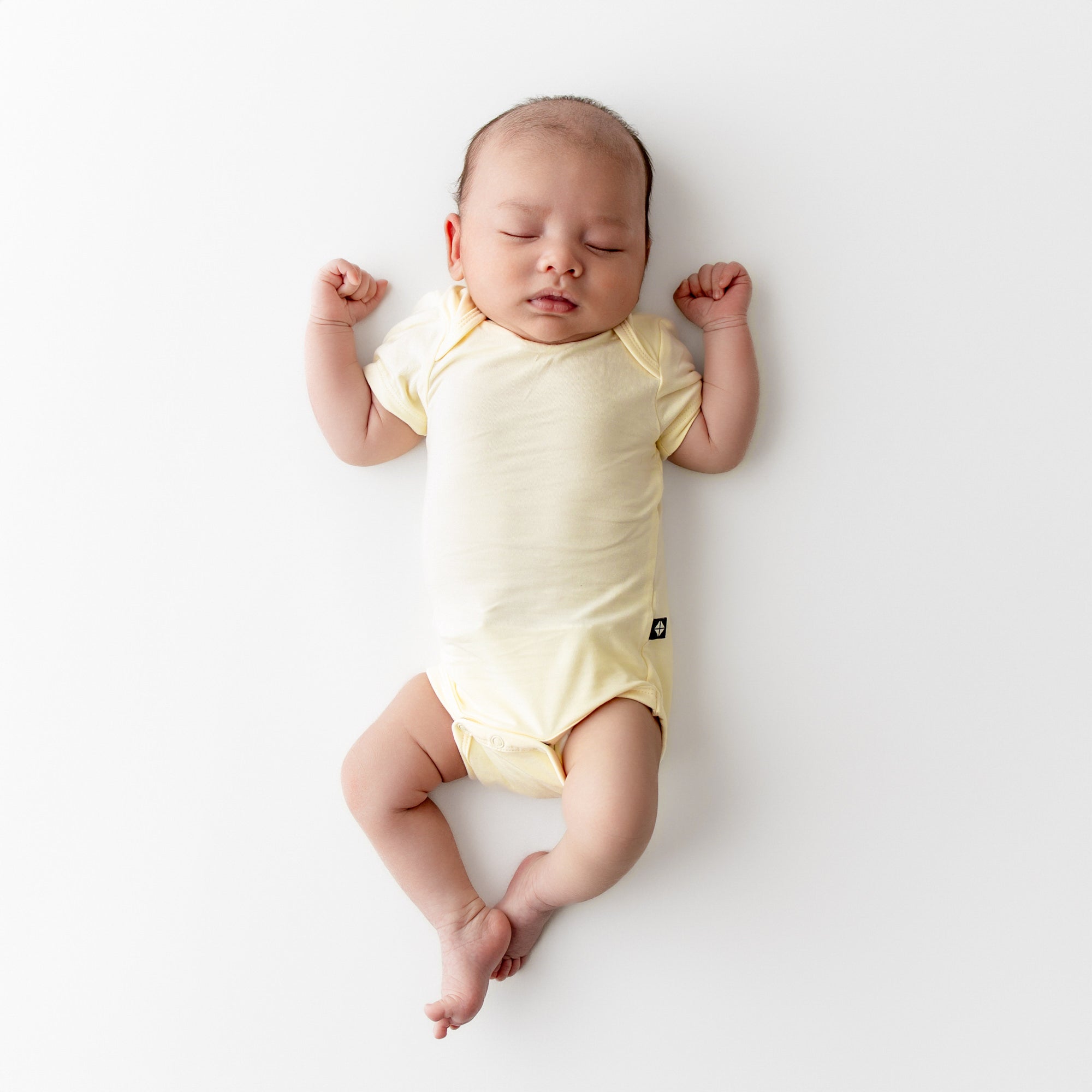Baby sleeping on a plain background wearing a pale yellow short sleeve bodysuit