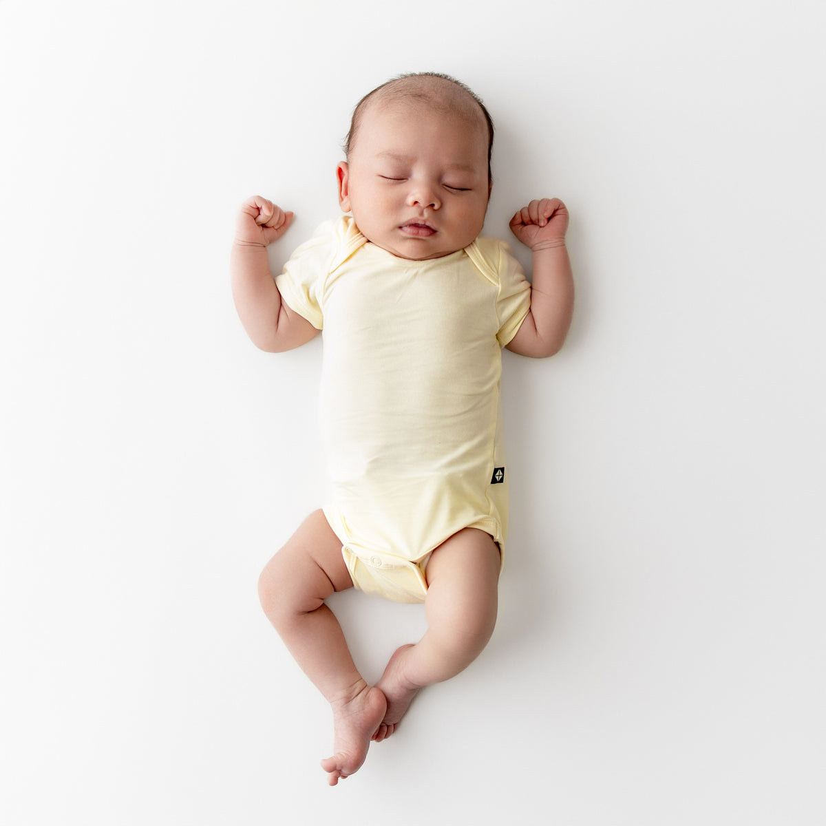 Baby sleeping on a plain background wearing a pale yellow short sleeve bodysuit