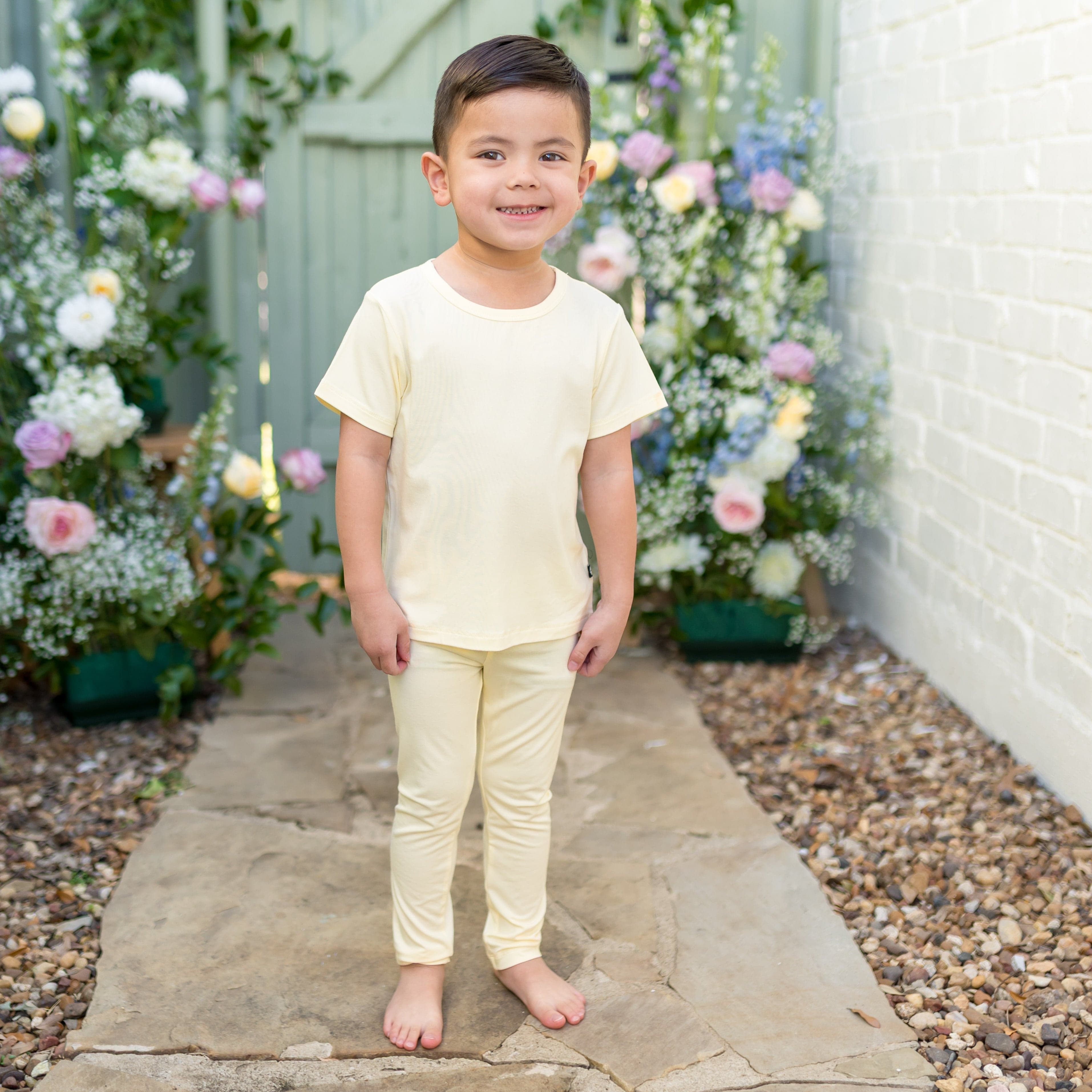 Child in a light yellow tee and matching leggings standing on a stone path with floral decorations.