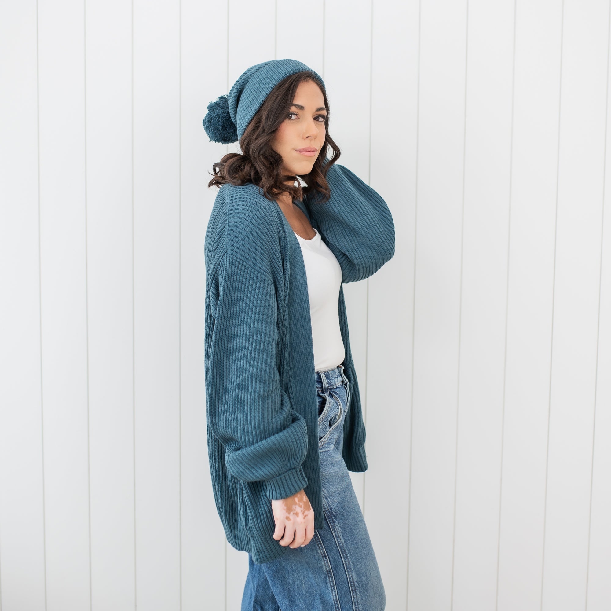 Female model standing in front of a white paneled wall wearing the Chunky Knit Women's Oversized Cardigan in Atlantic and matching chunky knit pom beanie