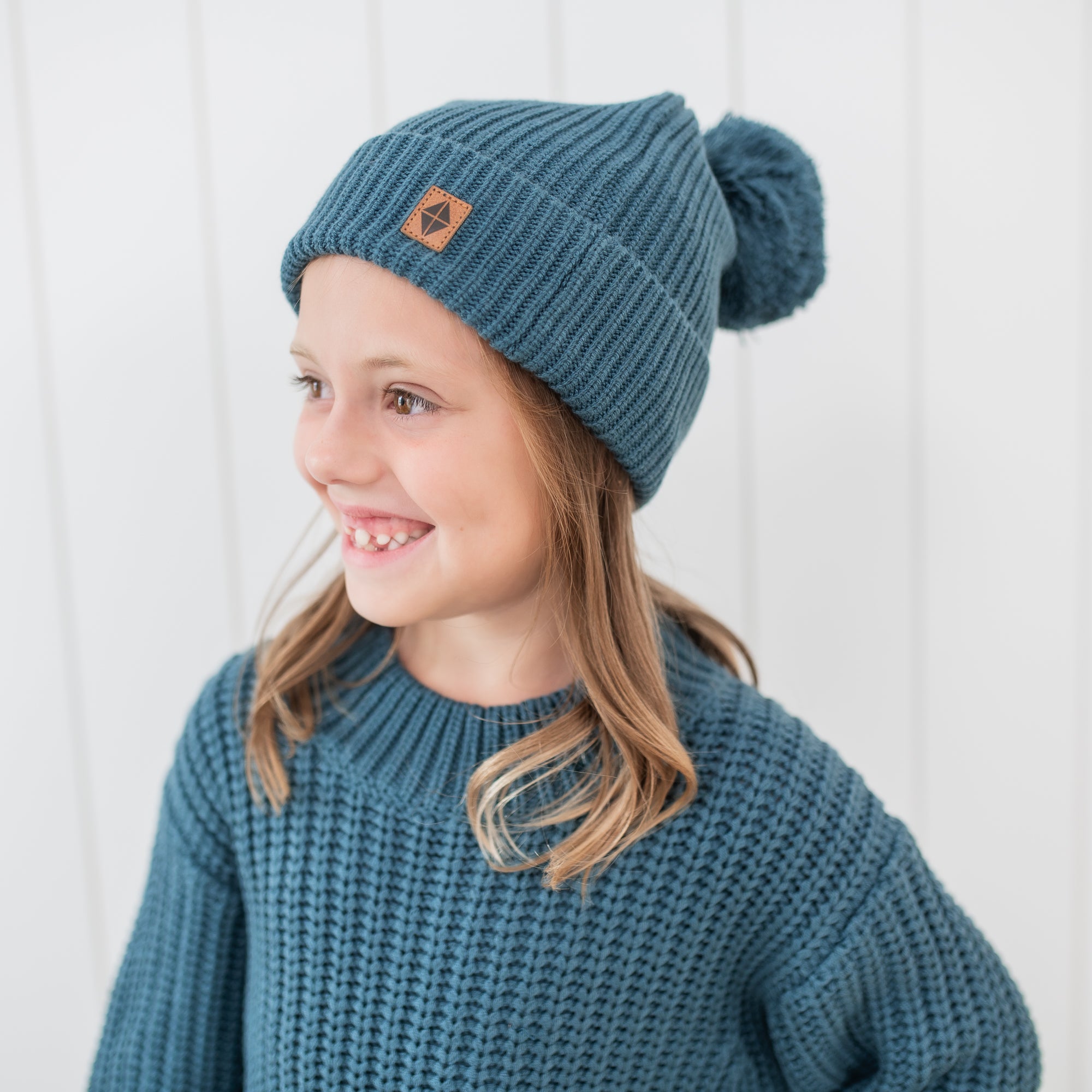 Close up of the Chunky Knit Pom Beanie in Atlantic on a young girl who is also wearing the Chunky Knit sweater in Atlantic