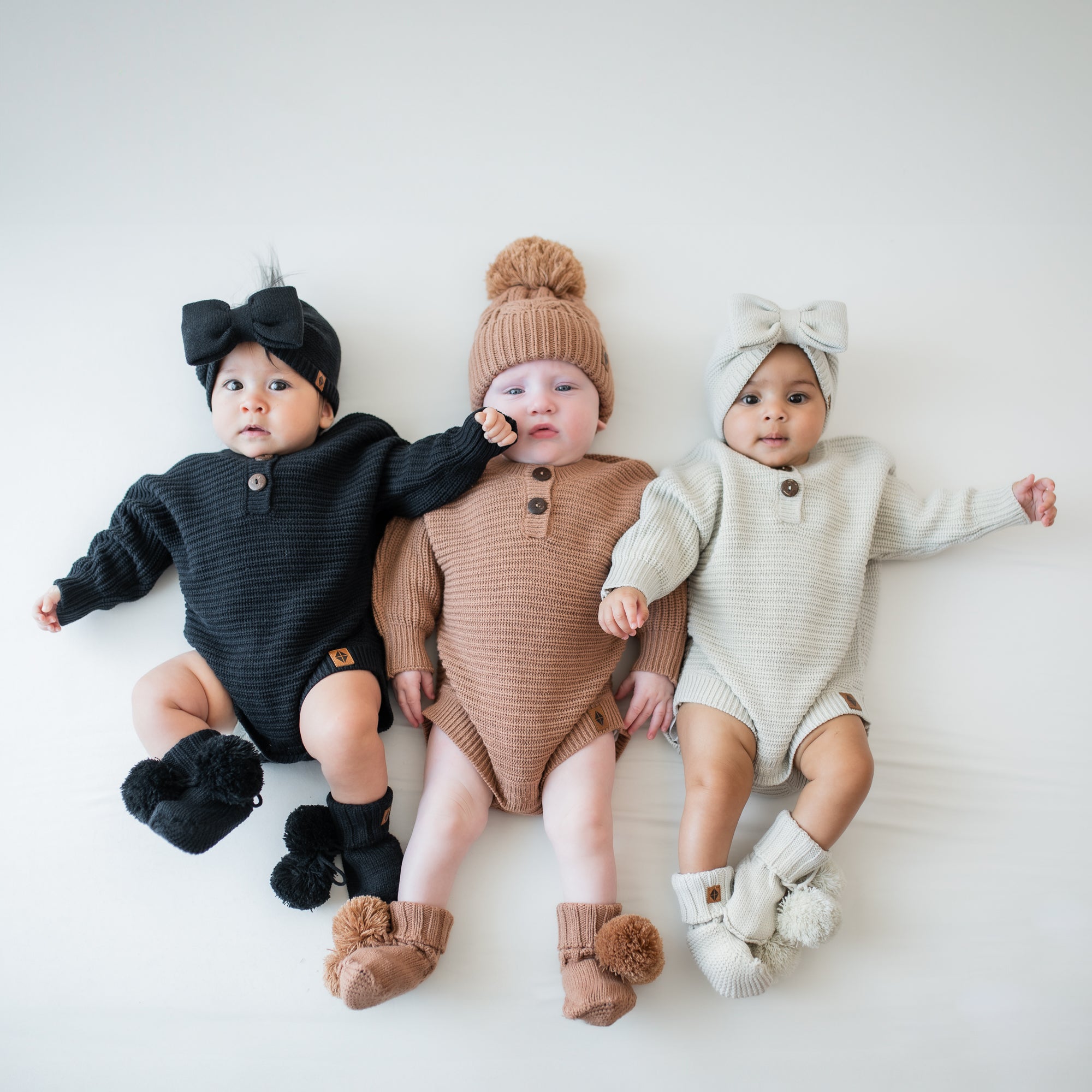 Three infants laying side by side wearing the Chunky Knit Romper in Latte, Oat and Midnight with matching knit booties
