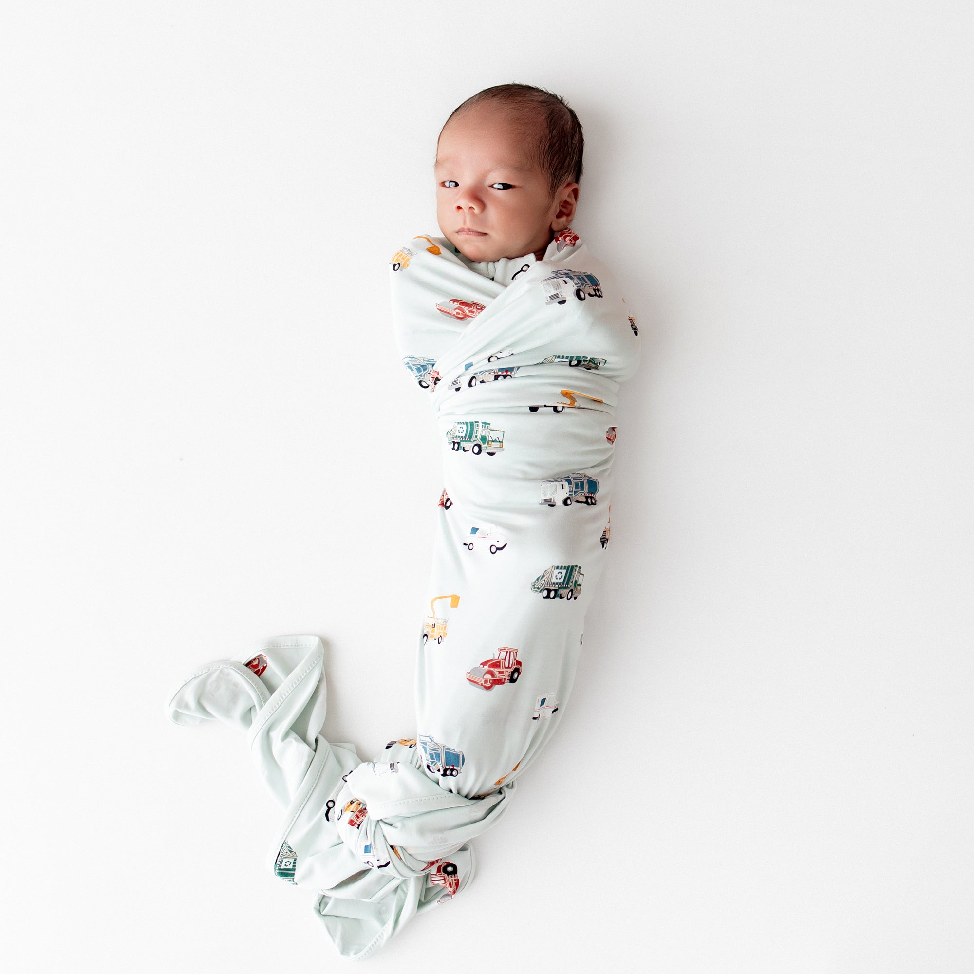 Newborn laying on a white surface swaddled in the soft and stretchy Swaddle Blanket in City Vehicles