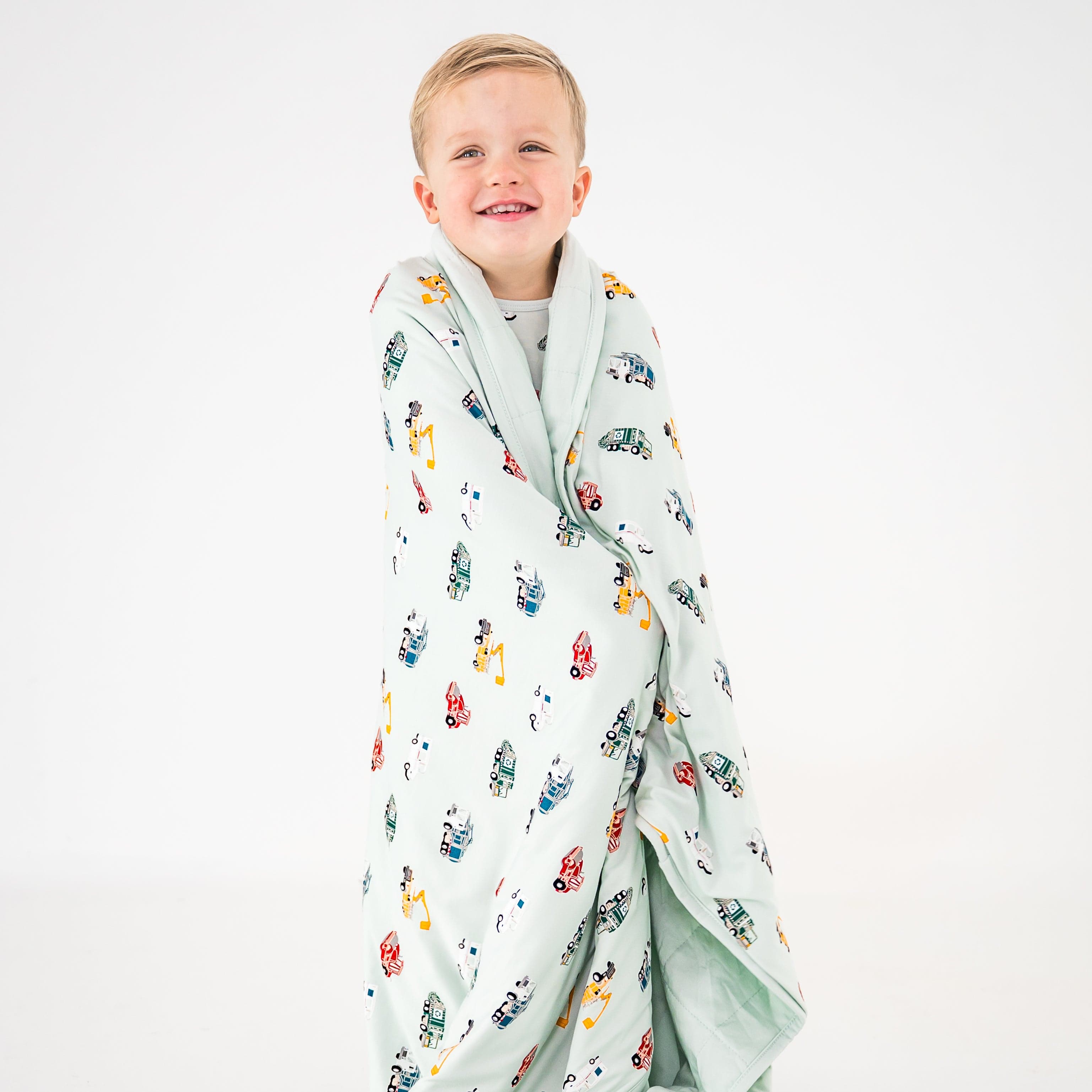 Young smiling boy standing in front of a white background with the Toddler Blanket in City Vehicles 1.0 around his shoulders