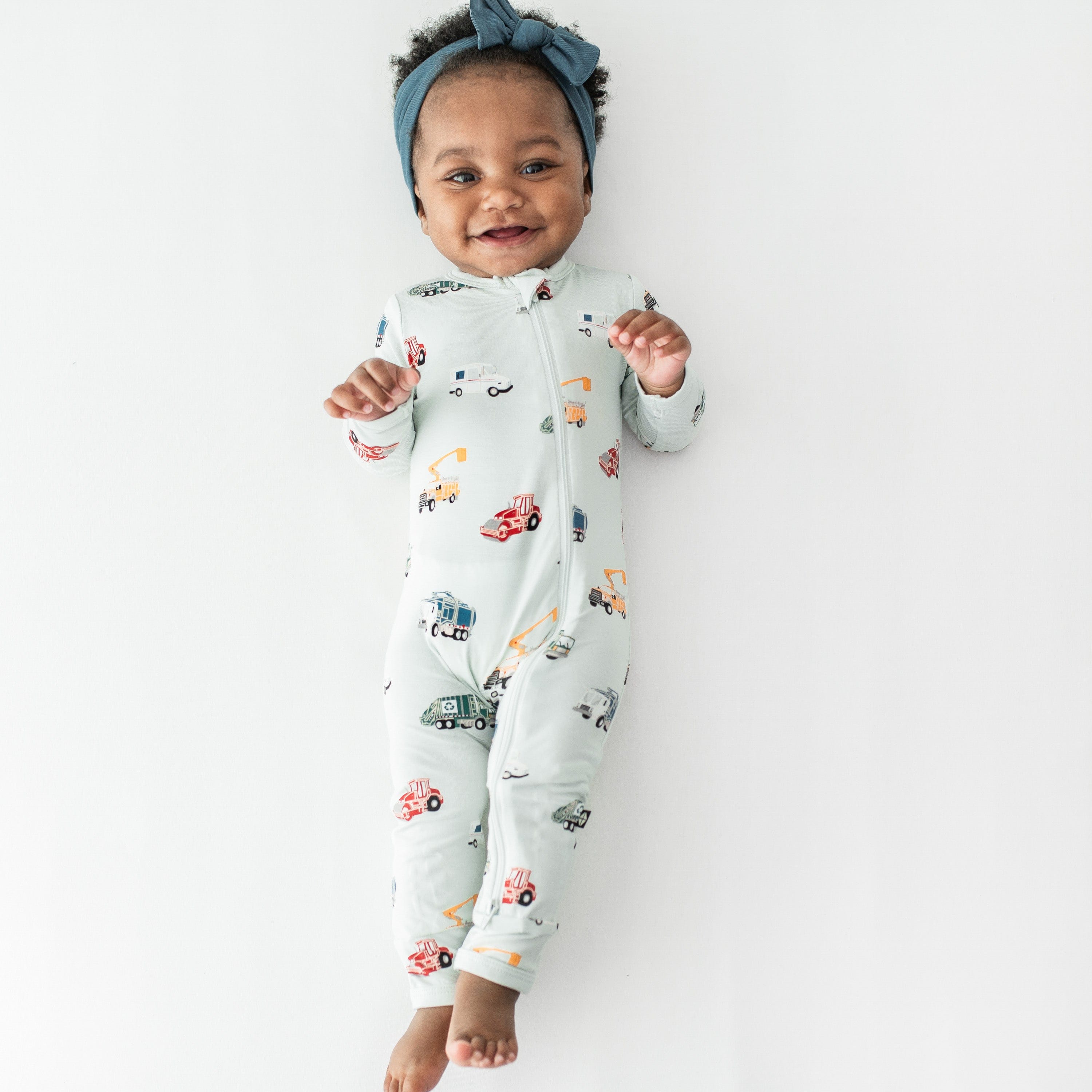 Young toddler girl laying stretched on a white surface wearing the Zippered Romper in City Vehicles and muted blue knotted bow headband