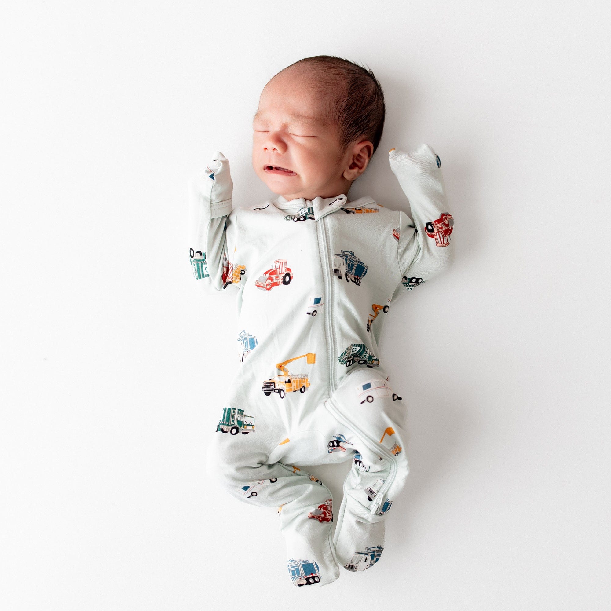 Newborn laying on a white surface wearing the Zippered Footie in City Vehicles with the fold over hand cuffs over the infants hands