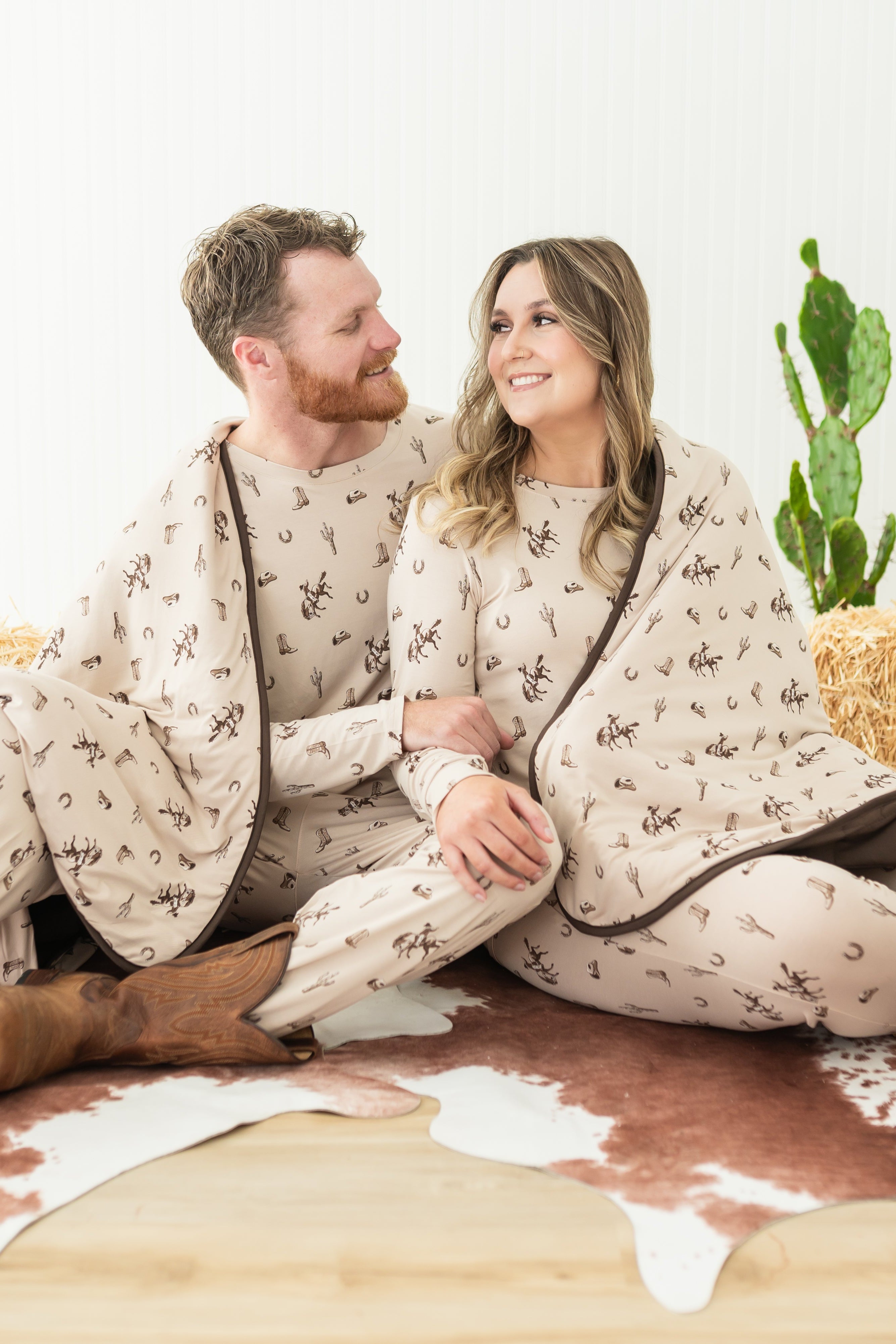 Couple sitting on the floor side by side matching in Classic Cowboy jogger sets with the matching adult blanket wrapped around them