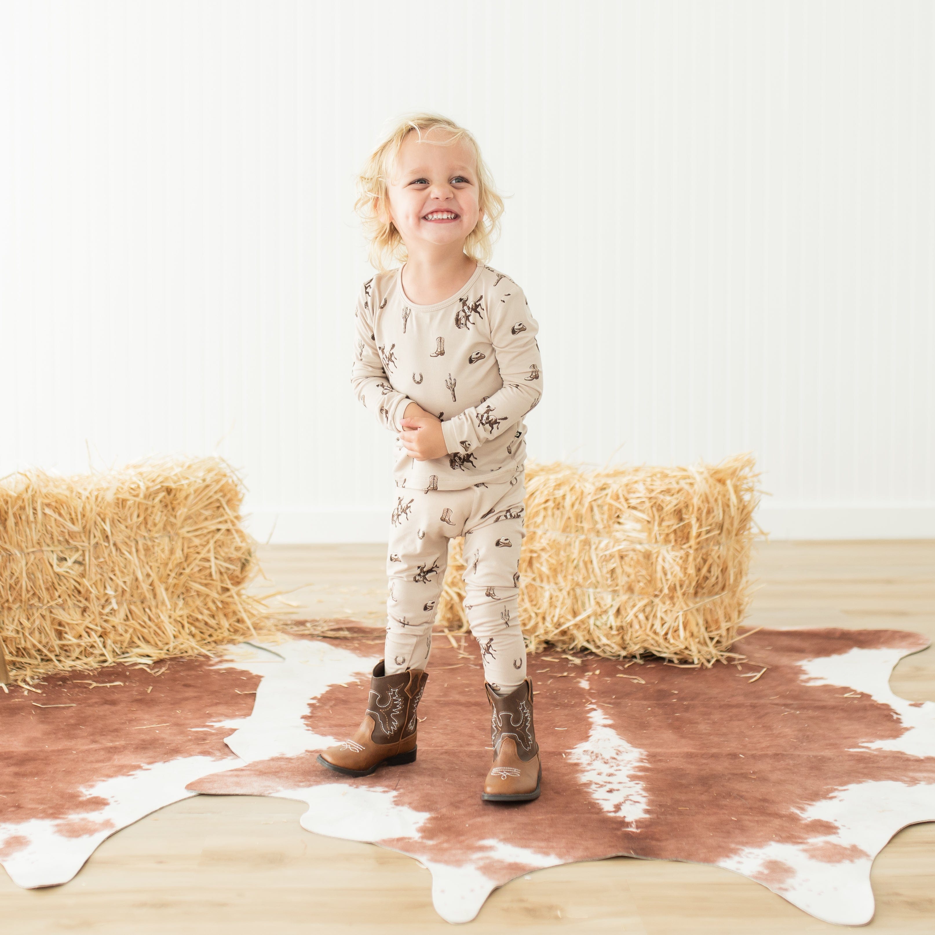 Toddler standing in front of hay bales wearing the Long Sleeve Pajamas in Classic Cowboy with cowboy boots