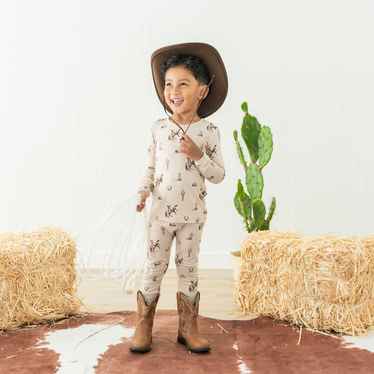 Young smiling boy wearing the Long Sleeve Pajamas in Classic cowboy with a cowboy hat and cowboy boots holding a white lasso in front of a cactus and hay bales