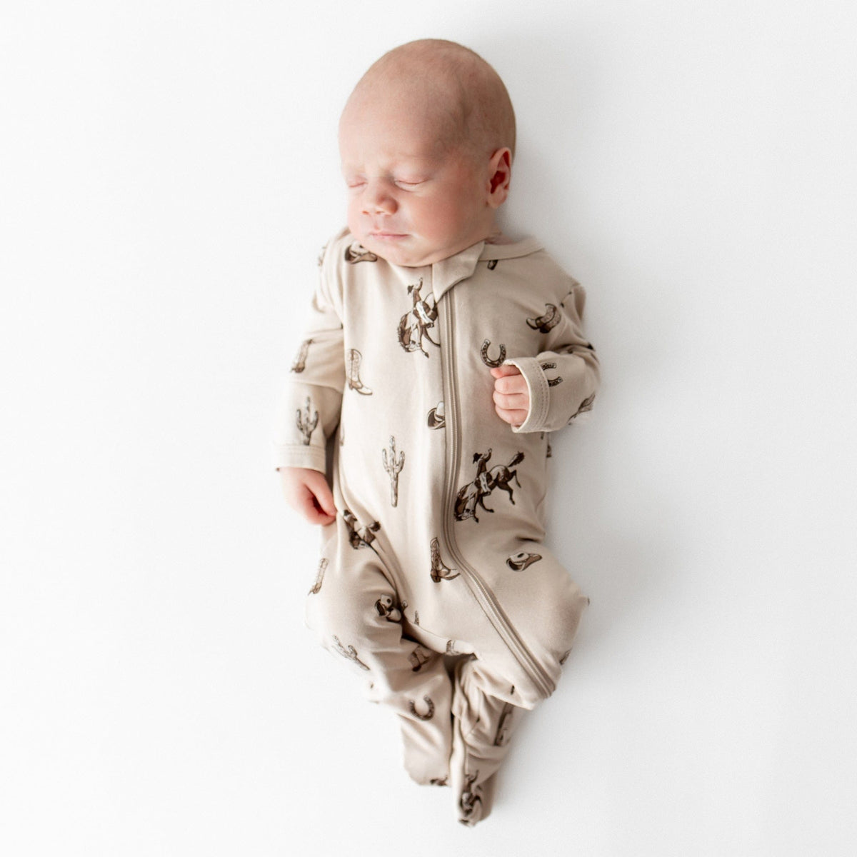 Sleeping newborn wearing the soft and breathable Zippered Footie in Classic Cowboy from Kyte Baby laying on a light cream surface