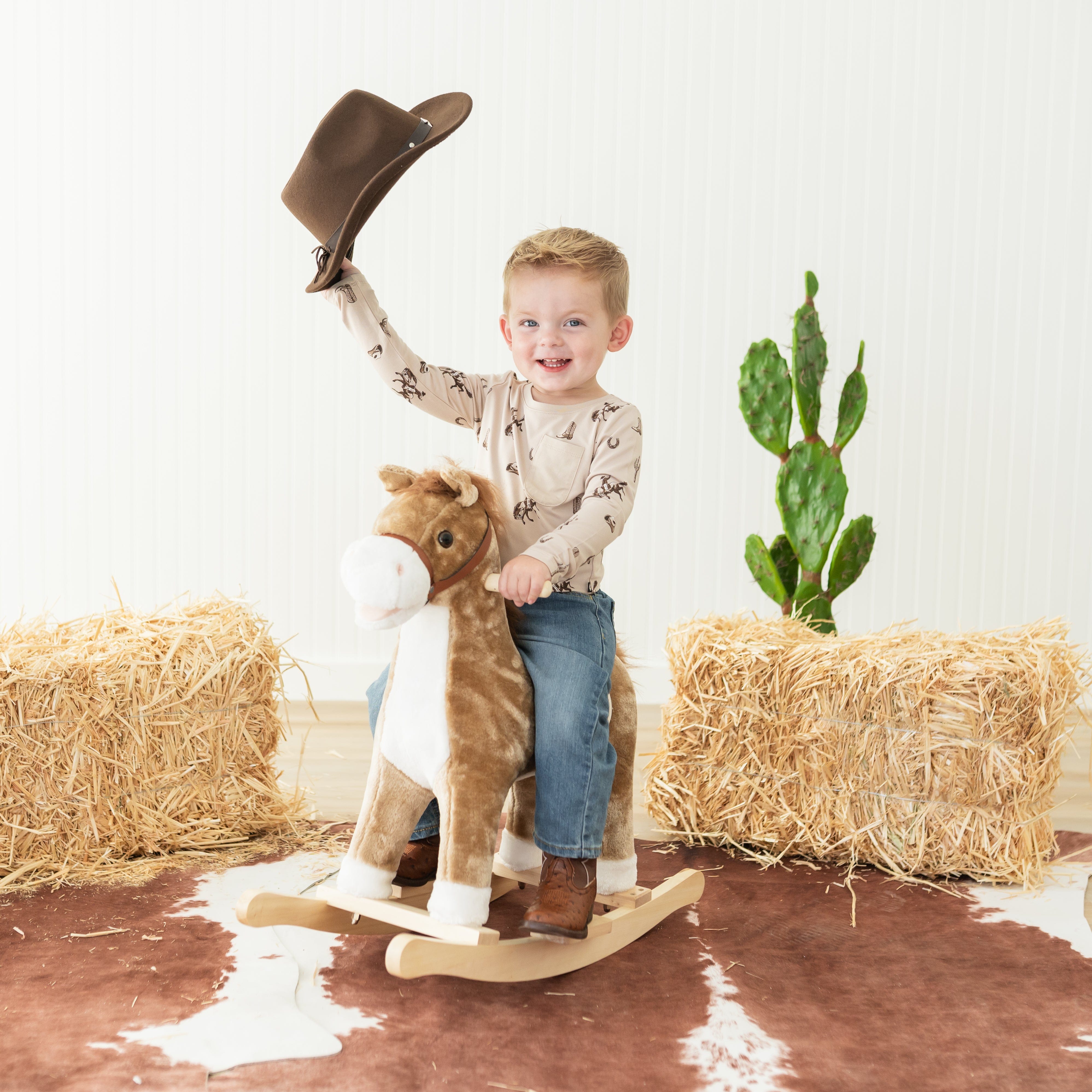 Young boy sitting on a rocking horse wearing the Long Sleeve Toddler Crew Neck Tee in Classic Cowboy with a cowboy hat in his hand in the air