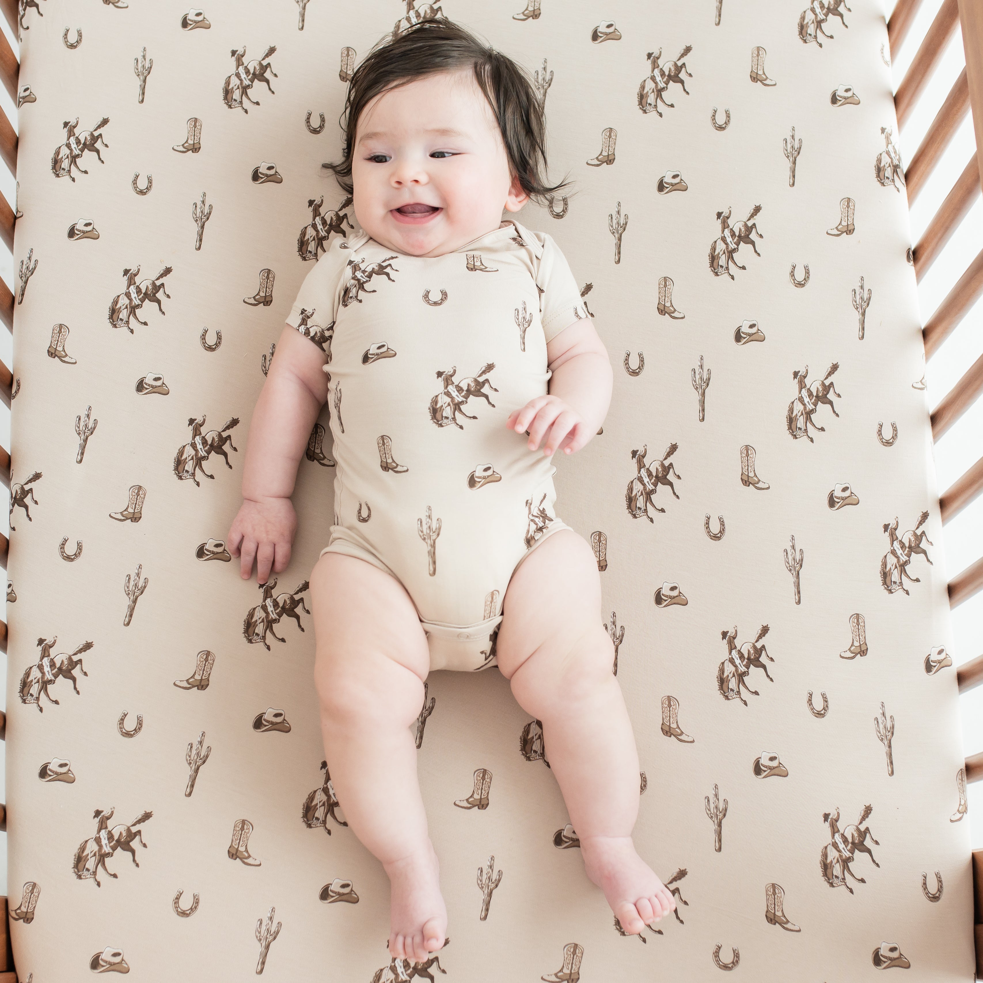 Smiling infant wearing the Bodysuit in Classic Cowboy laying in a crib on a matching crib sheet