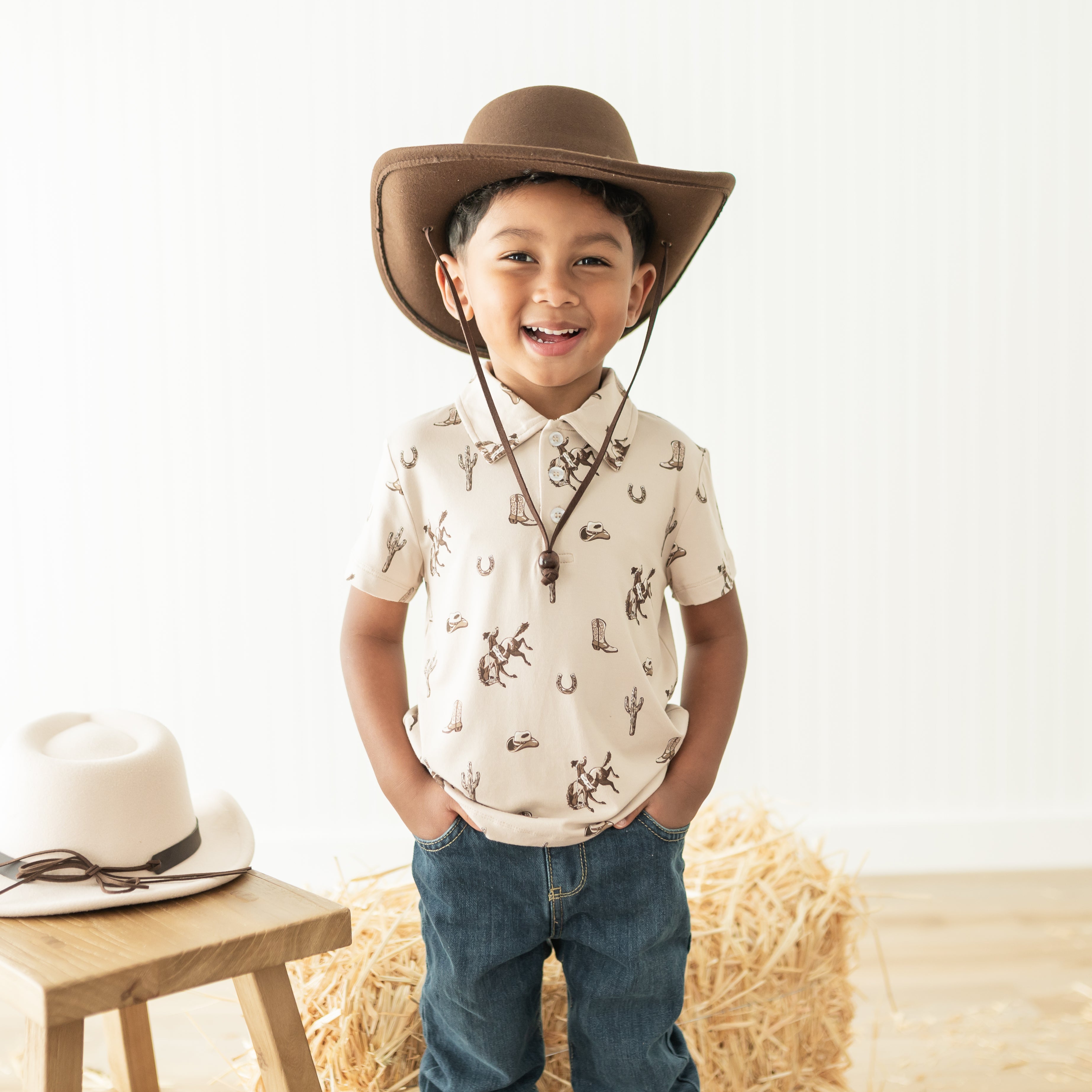 Young boy standing in front of hay bales wearing the Toddler Short Sleeve Polo in Classic Cowboy paired with a brown cowboy hat and medium wash jeans