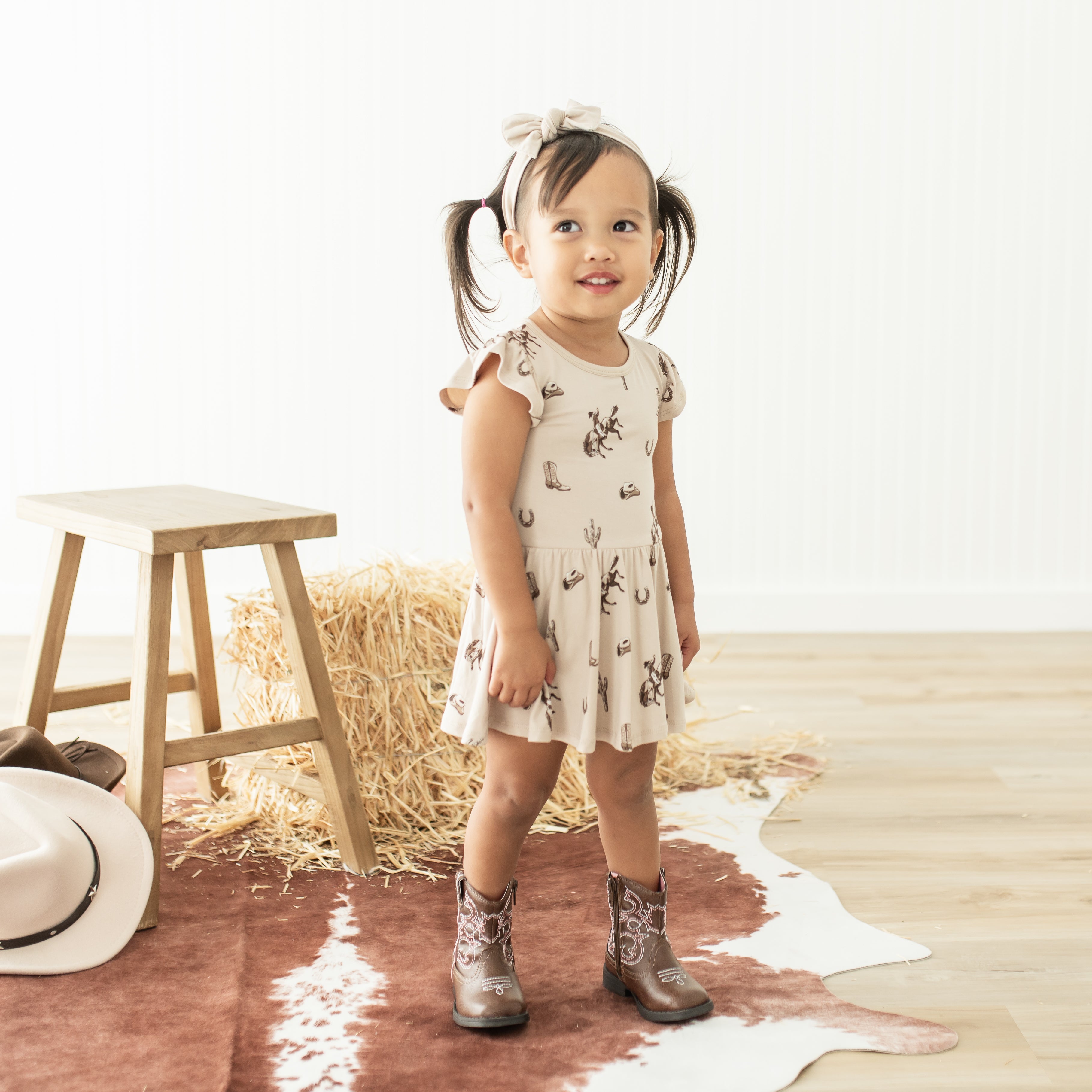 Young girl walking in front of western themed props wearing the Twirl Bodysuit Dress in Classic Cowboy with brown cowboy boots