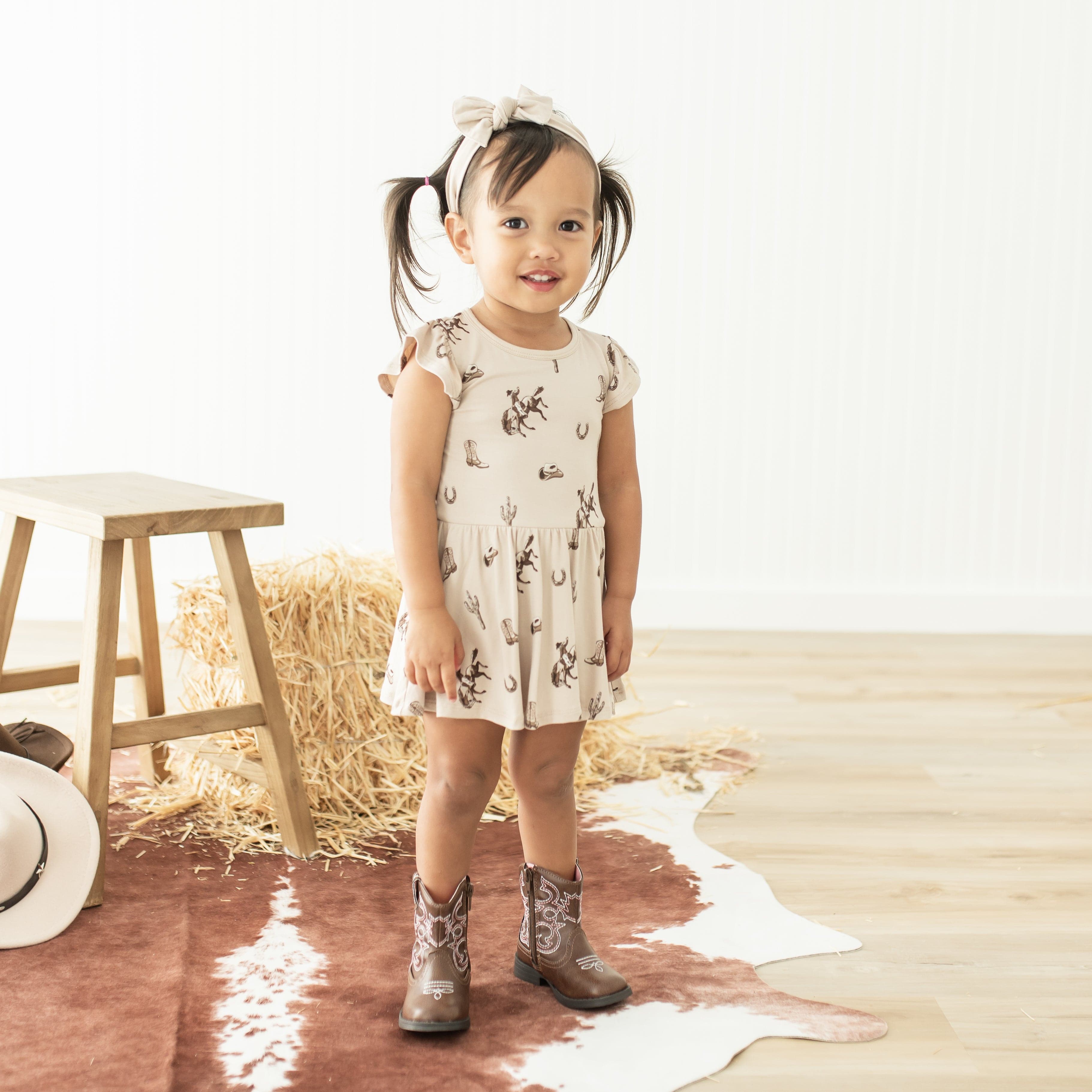 Young girl standing in front of a wooden stool and haybale wearing the Twirl Bodysuit Dress in Classic Cowboy with cowboy boots