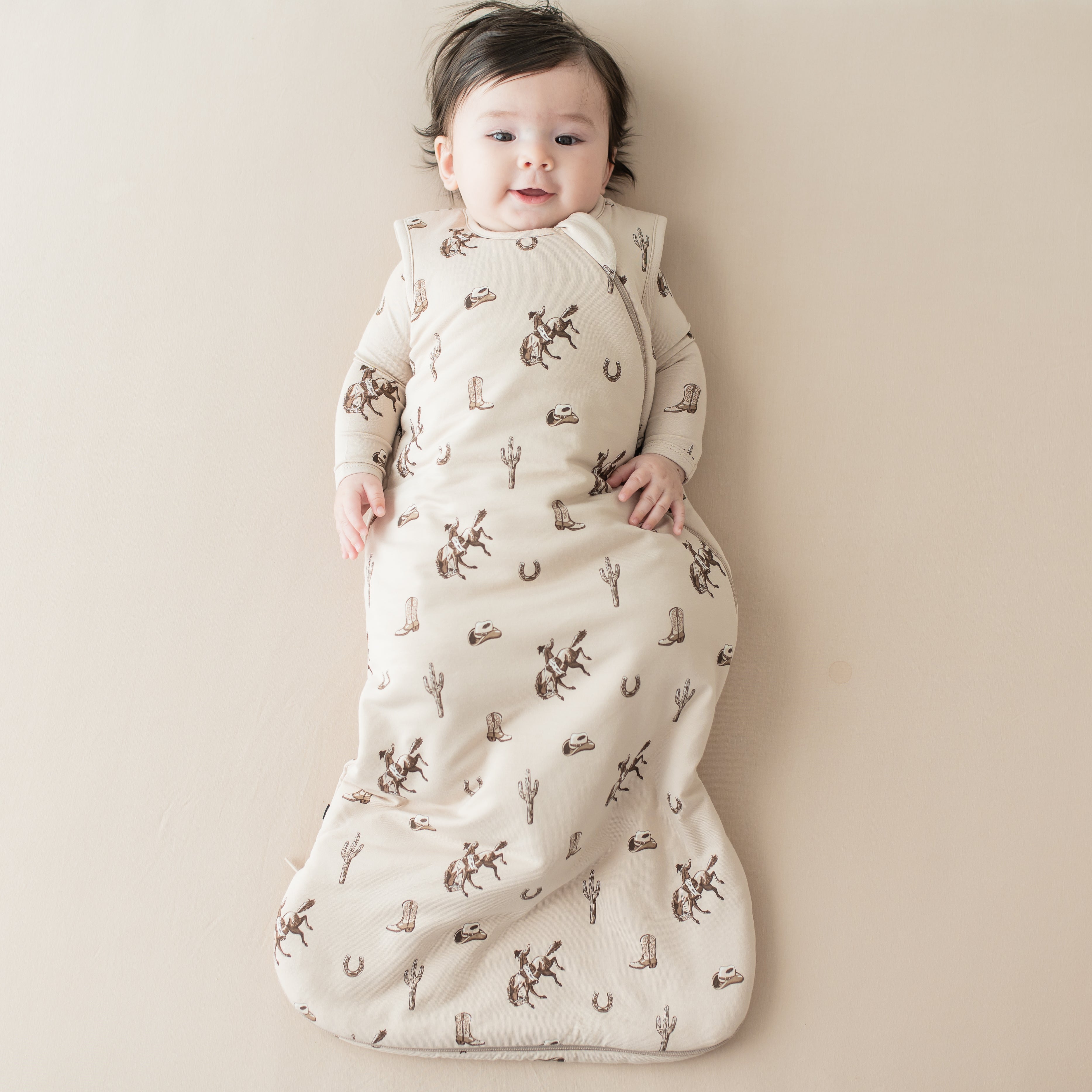Infant laying on a light beige surface wearing the Sleep Bag in Classic Cowboy 1.0 and matching long sleeve romper