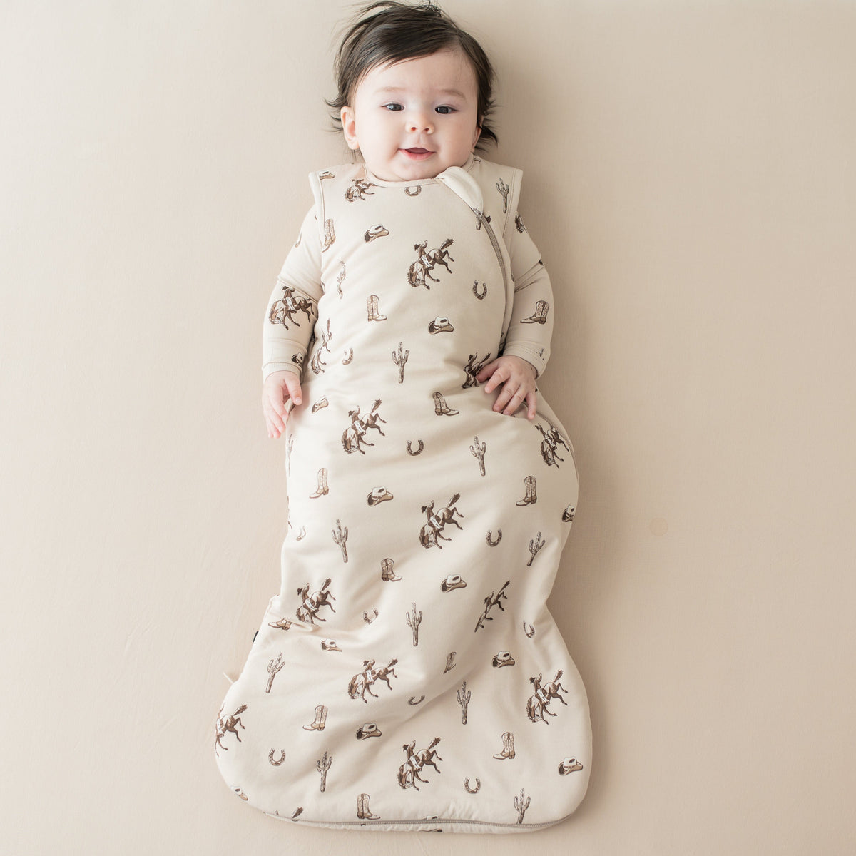 Infant laying on a light beige surface wearing the Sleep Bag in Classic Cowboy 1.0 and matching long sleeve romper