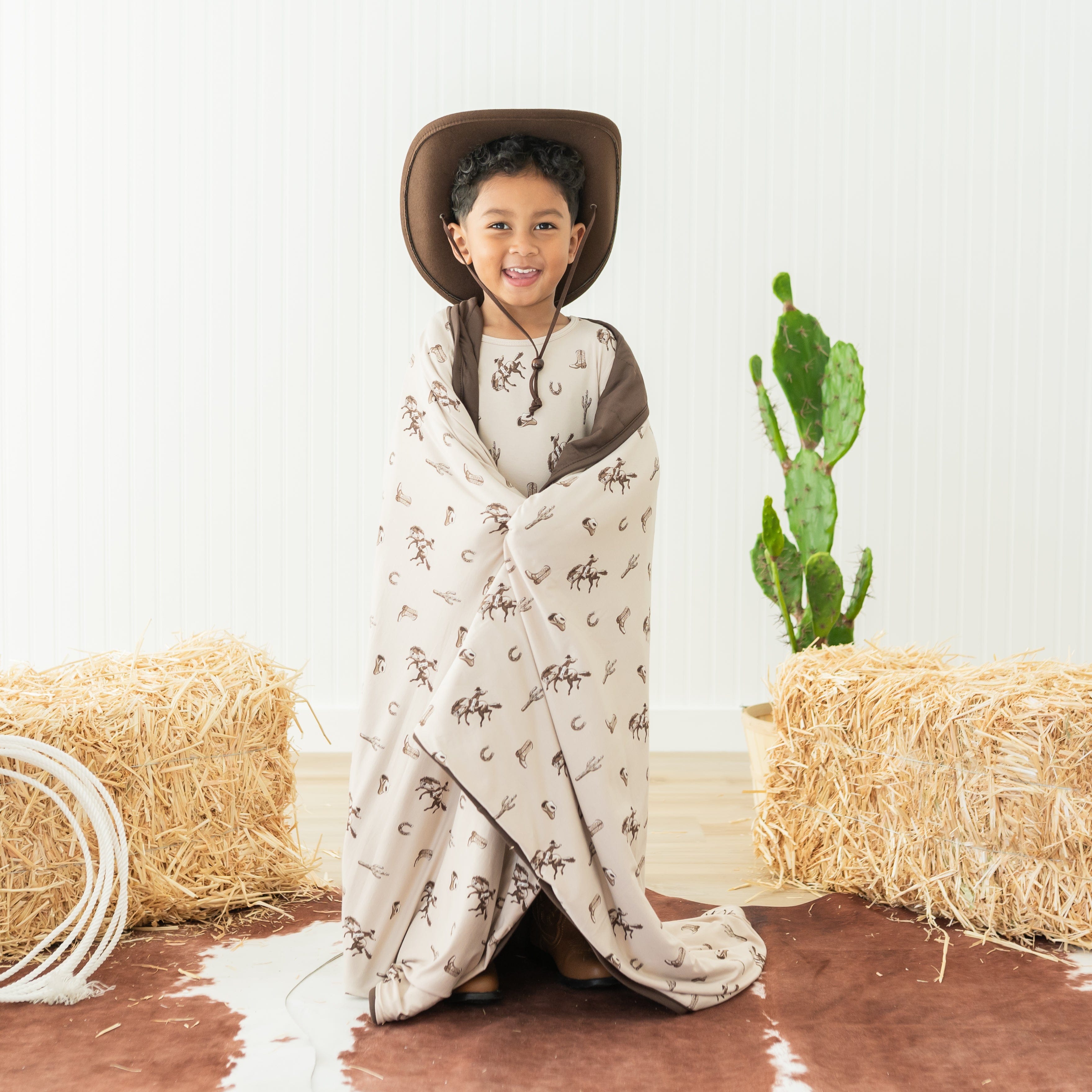 Young boy standing in front of a western inspired back drop wearing a cowboy hat with the Toddler Blanket in Classic Cowboy 1.0 wrapped around him