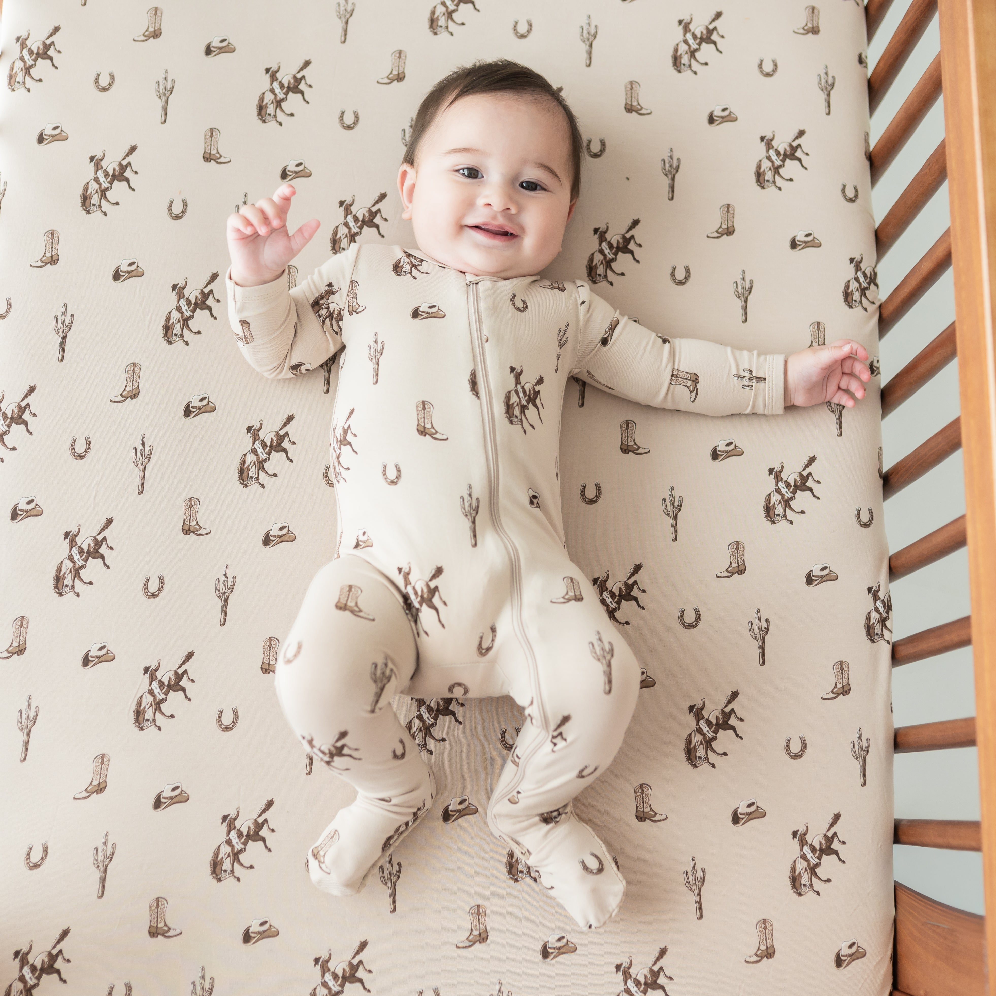 Infant wearing the Zippered Footie in Classic Cowboy laying in a crib on a matching crib sheet
