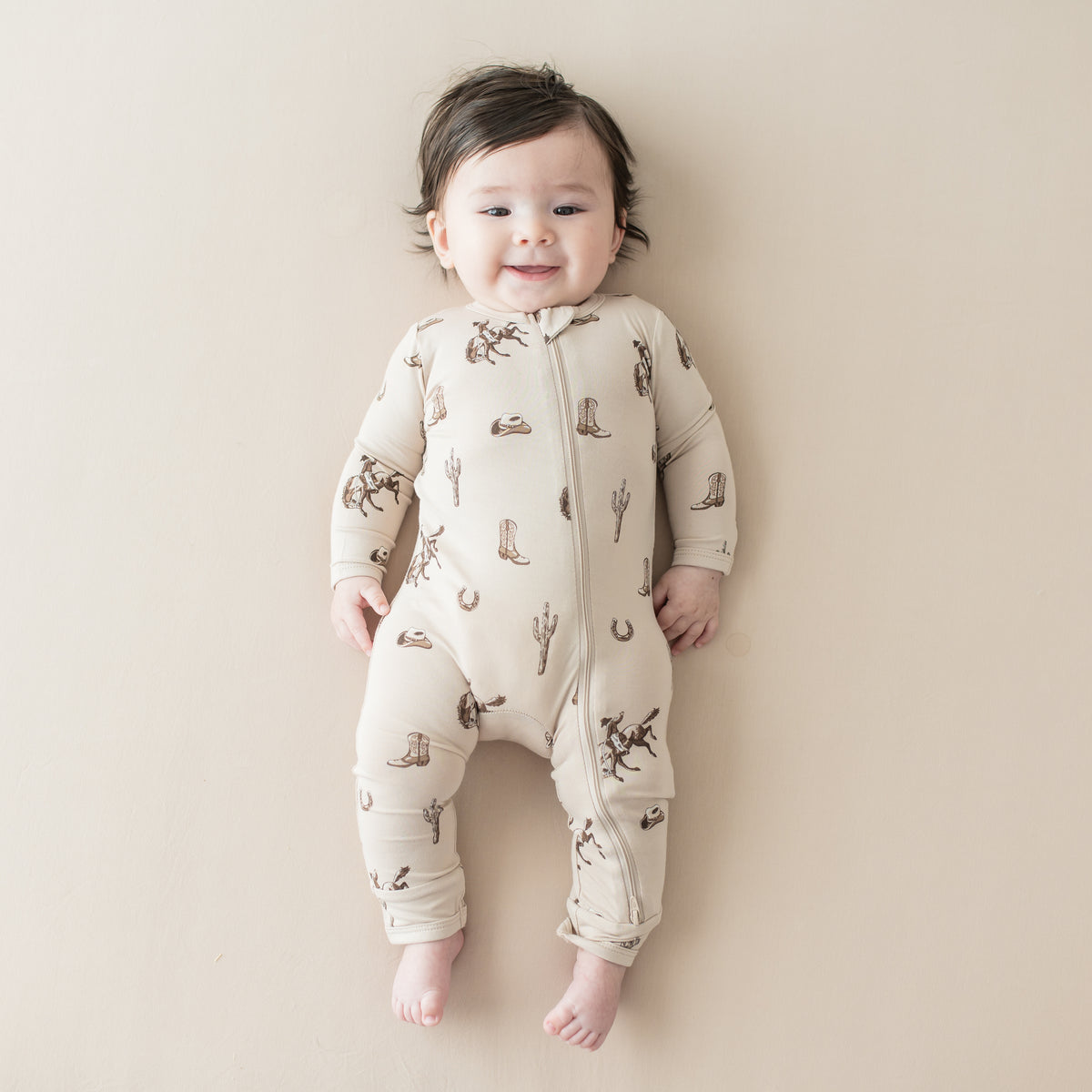 Infant laying on a light neutral surface wearing the Zippered Romper in Classic Cowboy