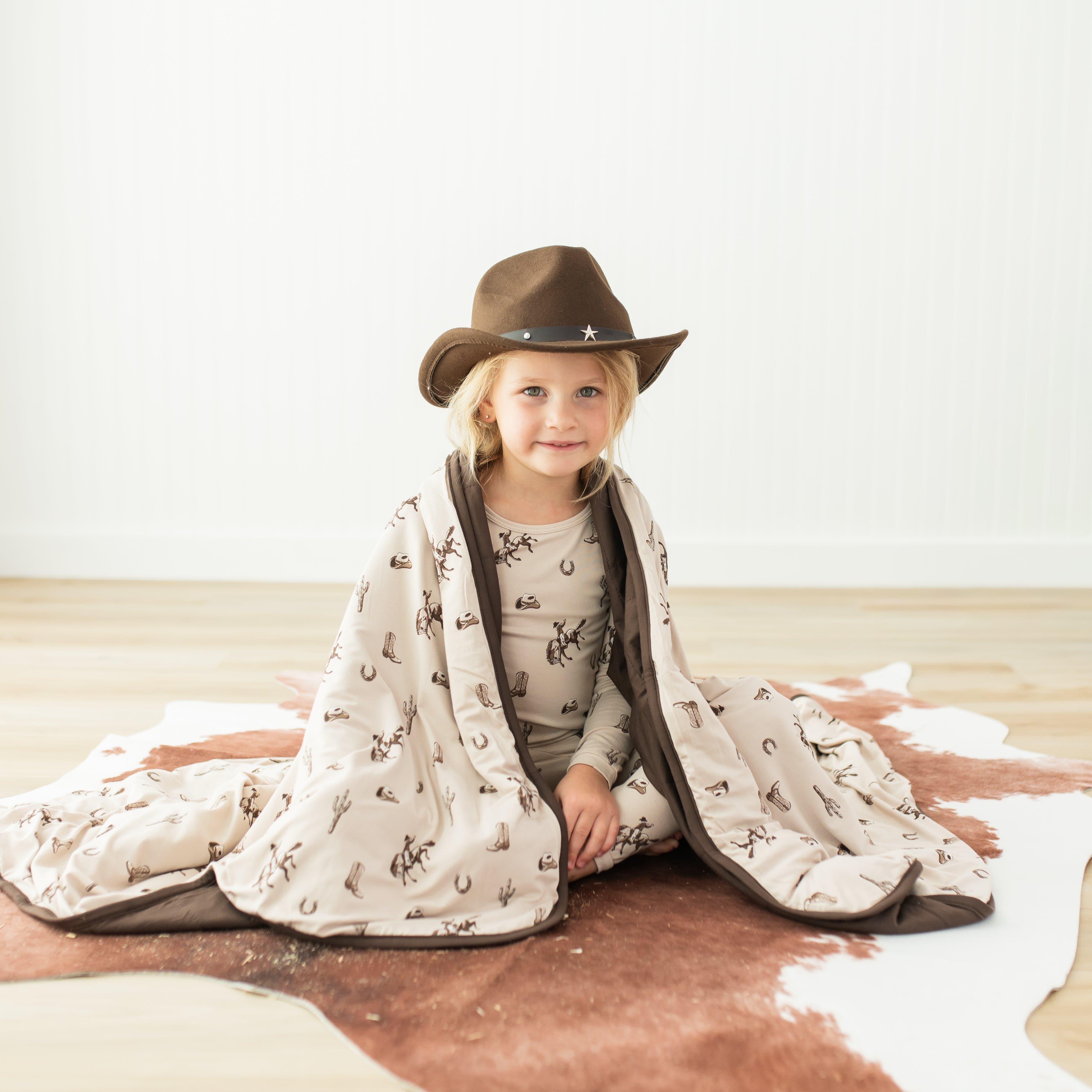 Young child sitting on the floor with the Youth Blanket in Classic Cowboy 2.5 wrapped around their shoulders wearing matching long sleeve pajamas and a brown cowboy hat