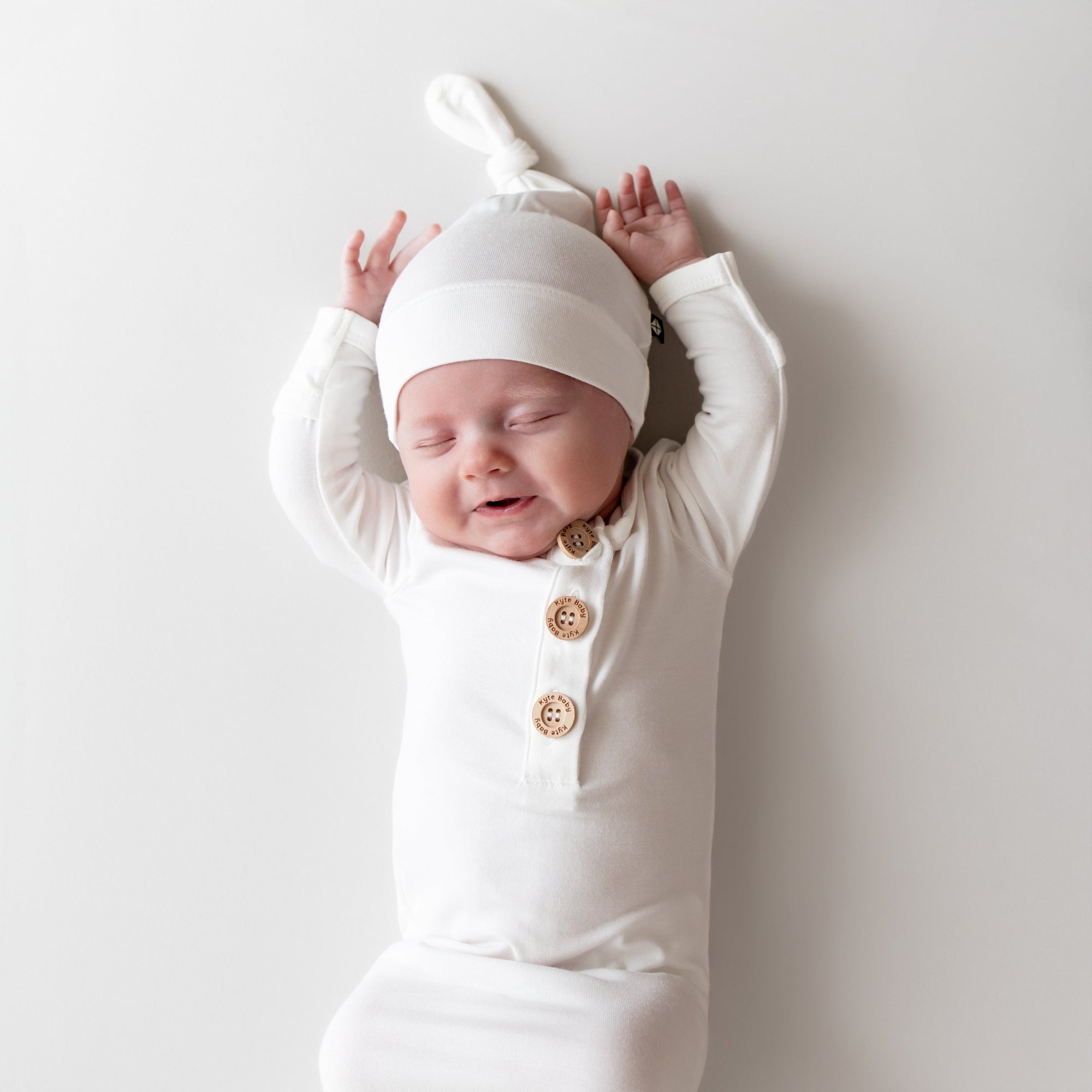 Newborn baby in a white outfit with buttons, wearing a matching white hat, against a plain background.