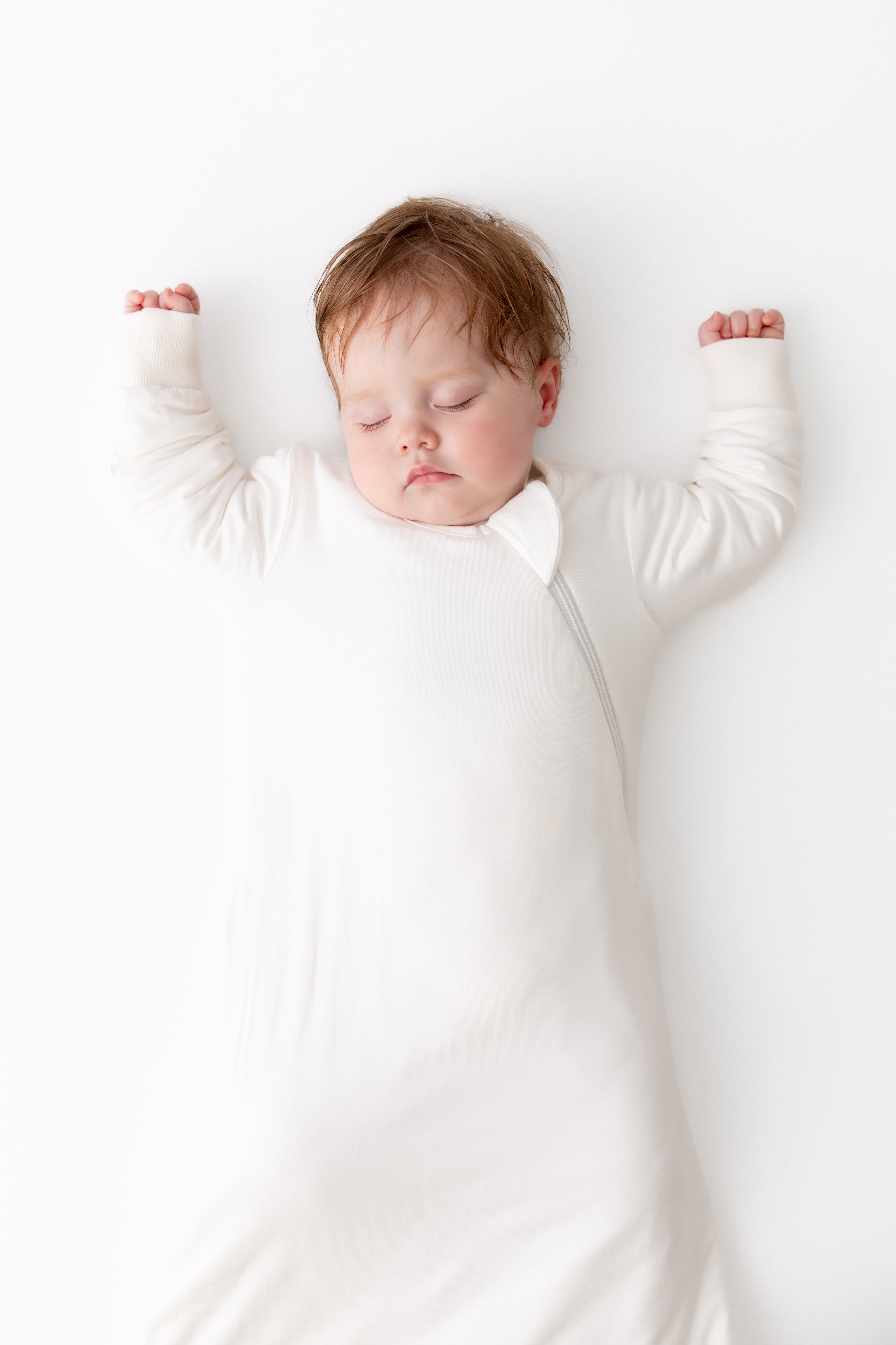 A sleeping newborn baby wearing a long-sleeve white sleep sack, lying flat on a clean white background with arms raised.