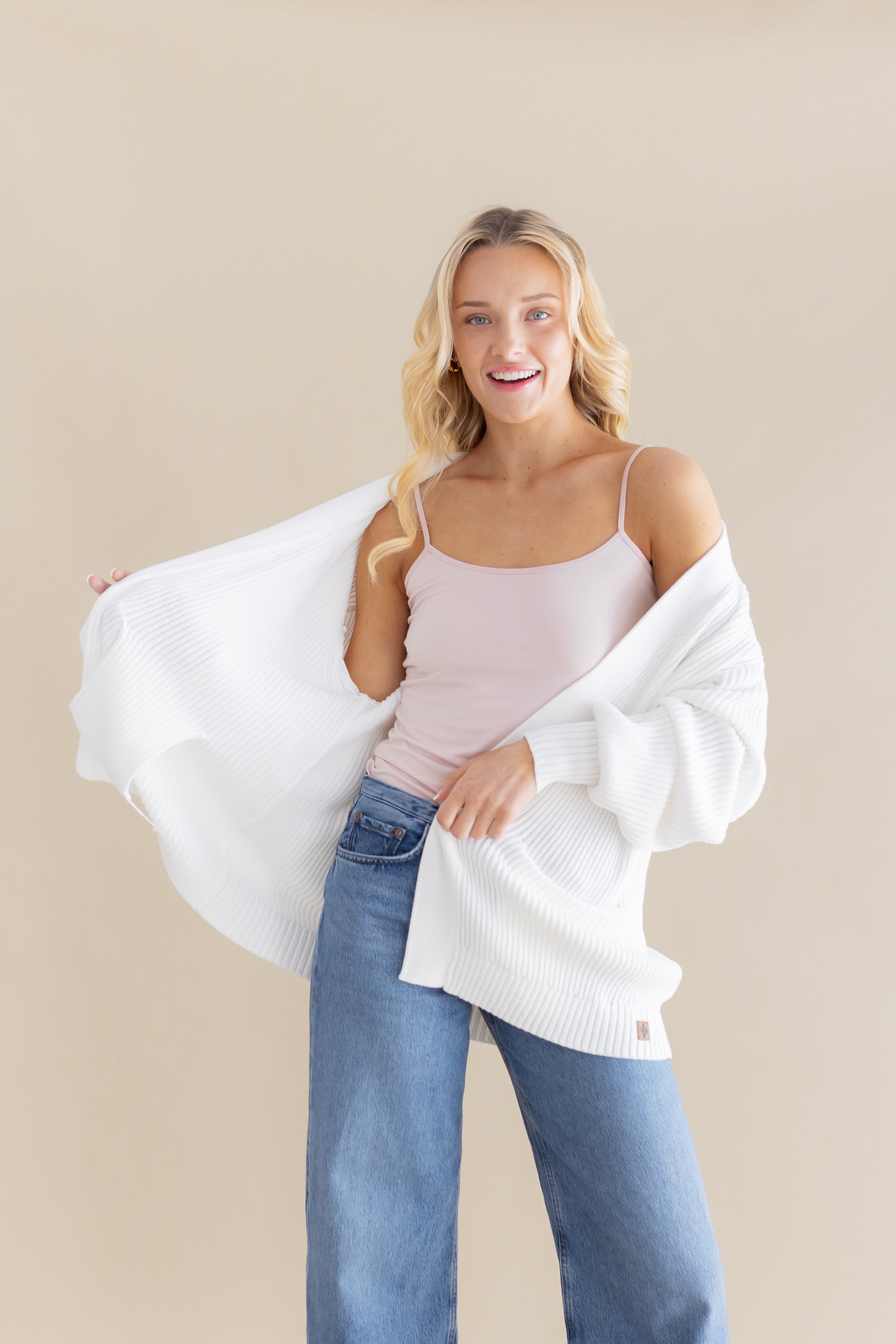 Female model posing wearing the Chunky Knit Women's Oversized Cardigan in Cloud around her shoulders paired with a light pink camisole and light wash jeans