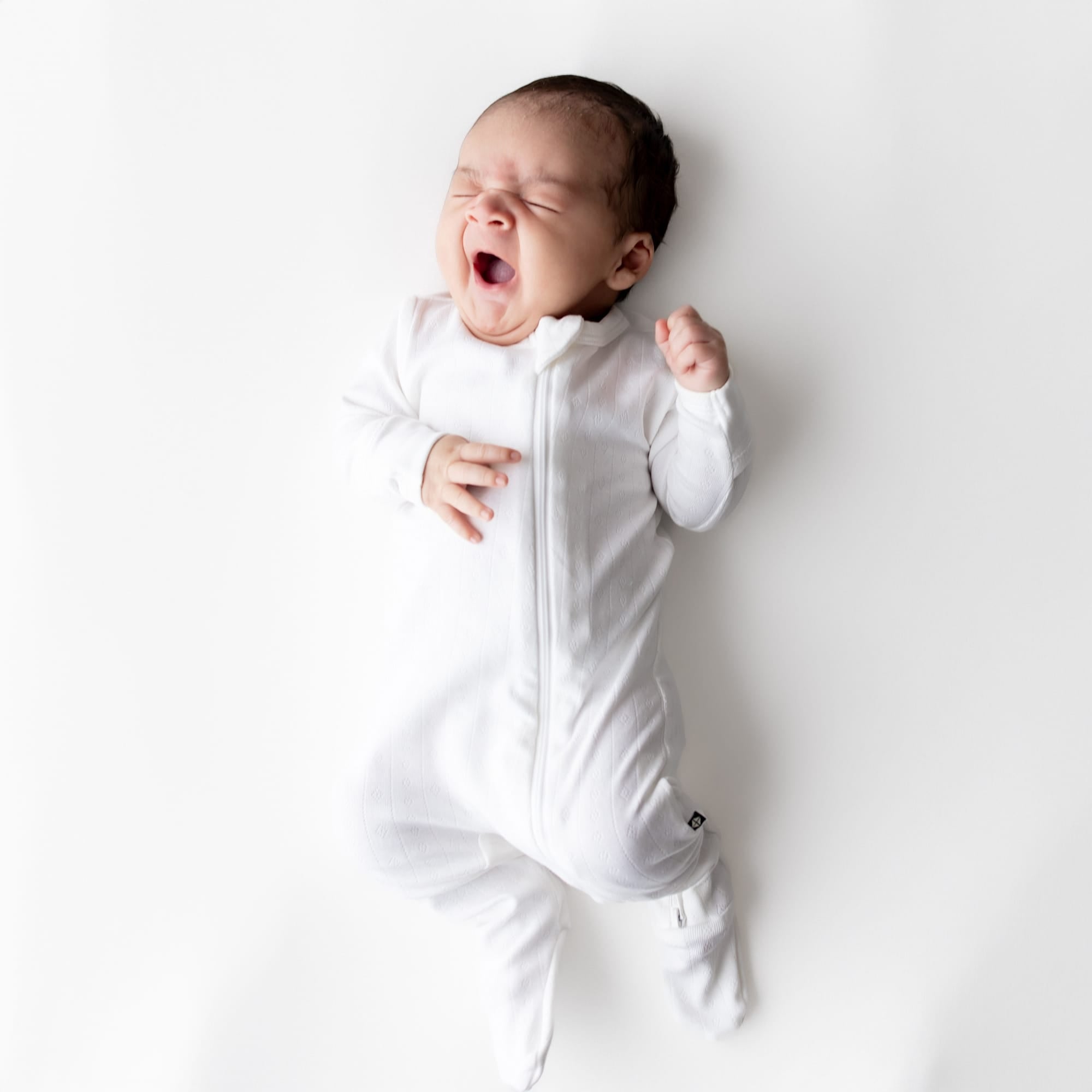 Yawning newborn wearing the Pointelle Zippered Footie in Cloud on a white surface
