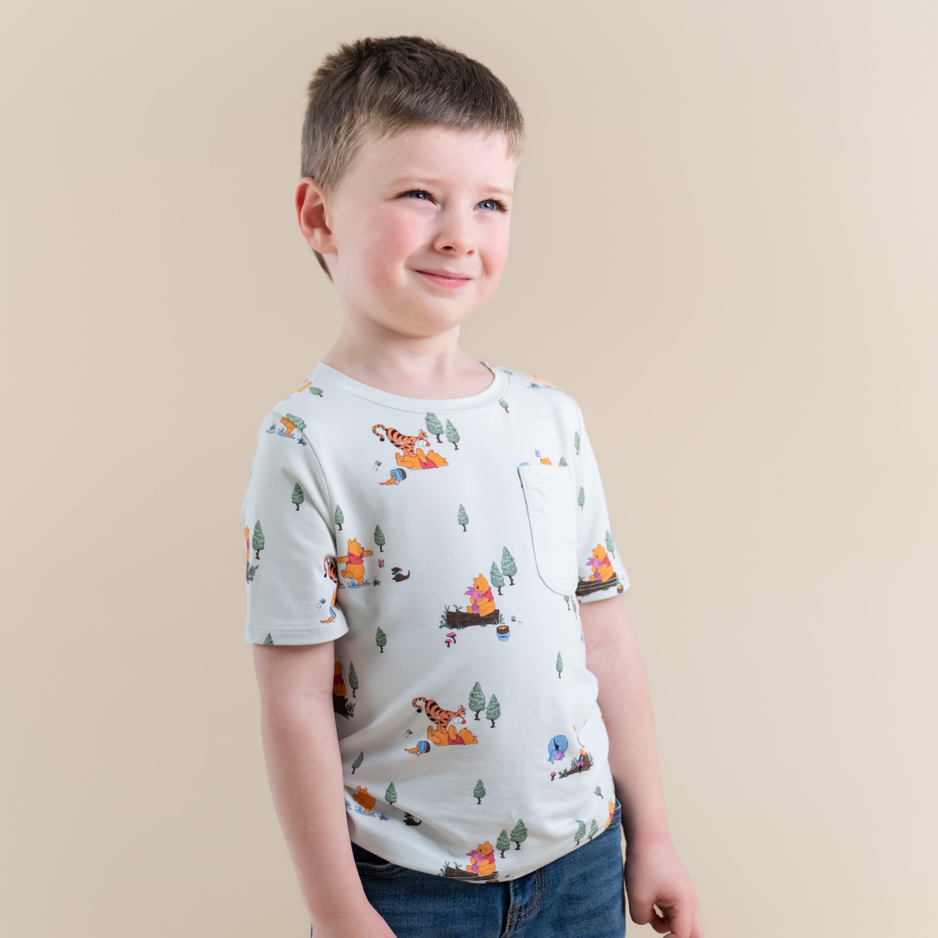 Smiling boy looking off to the side wearing the Toddler Crew Neck Tee in Winnie the Pooh Hundred Acre Wood