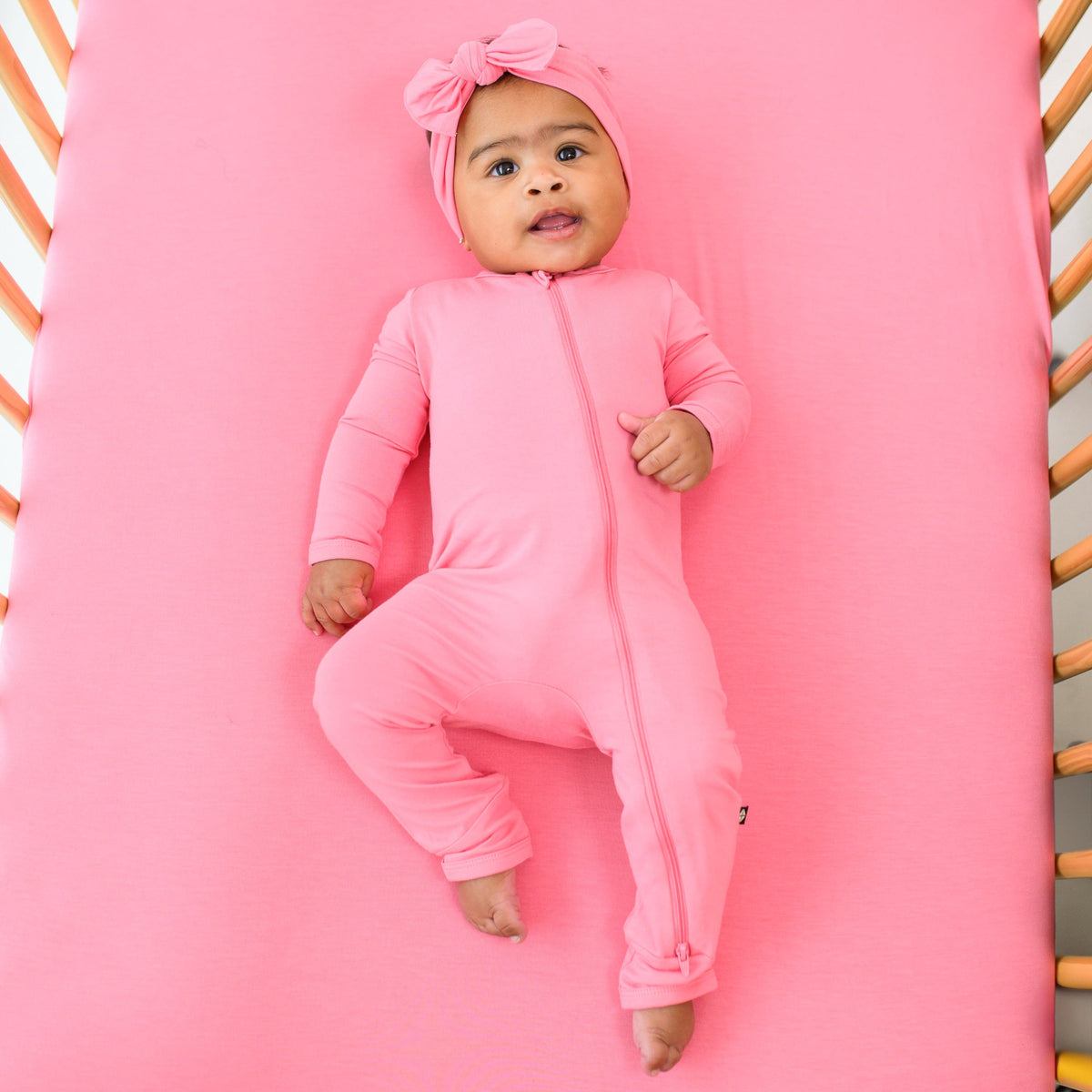 Infant wearing Kyte Baby Zippered Romper in Guava laying on matching Crib Sheet