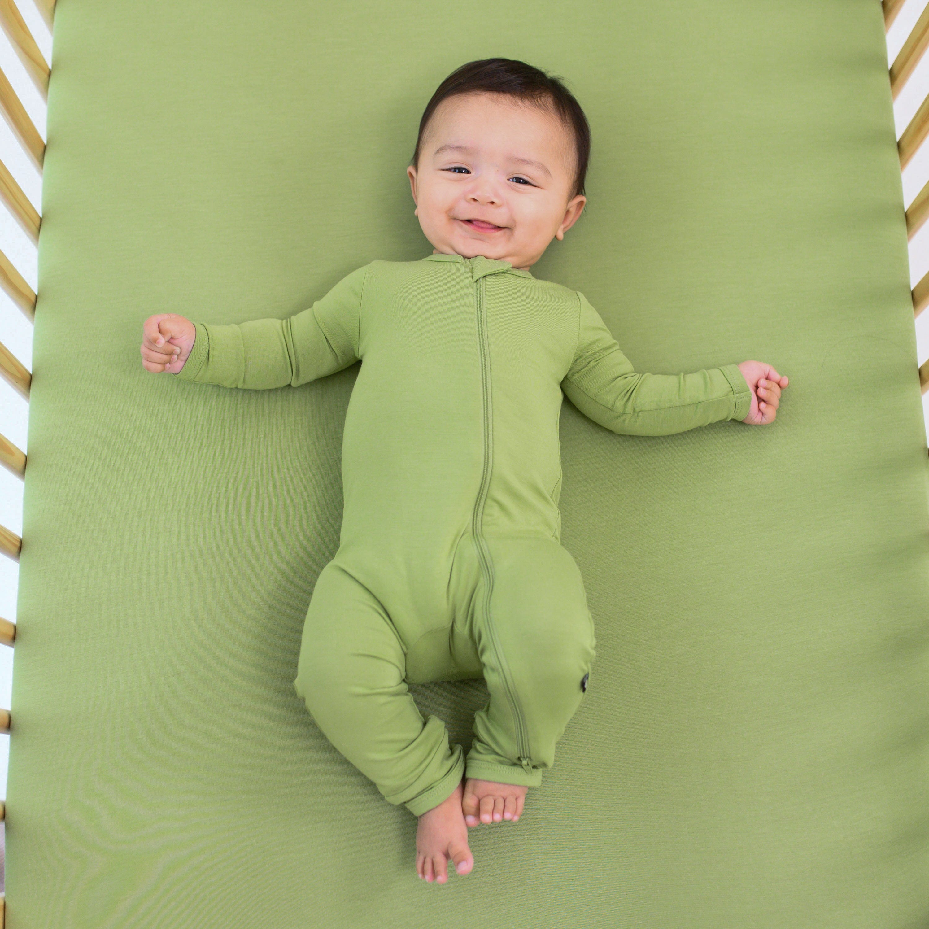 Infant wearing Kyte Baby Zippered Romper in Honu green laying on matching Crib Sheet