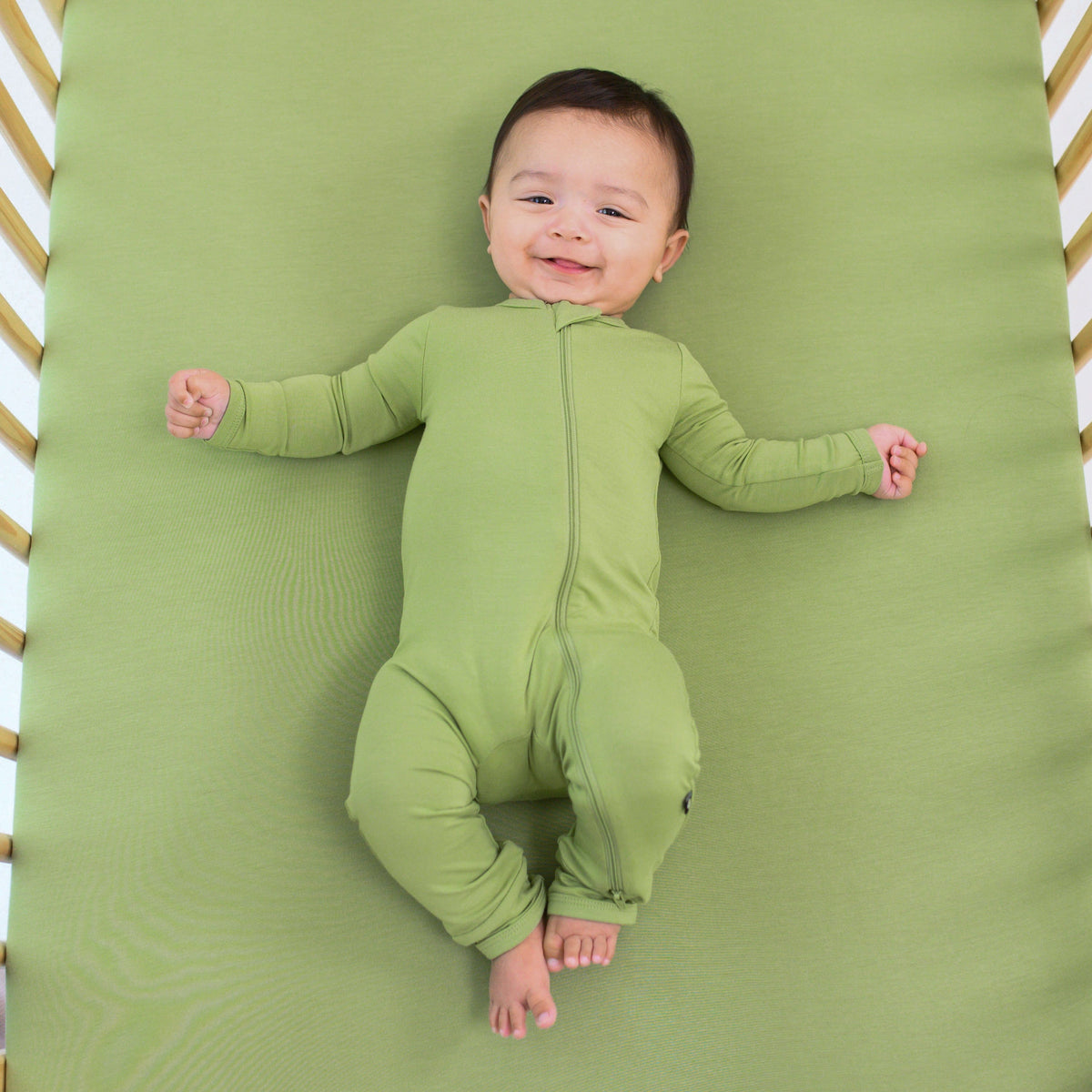 Infant wearing Kyte Baby Zippered Romper in Honu green laying on matching Crib Sheet