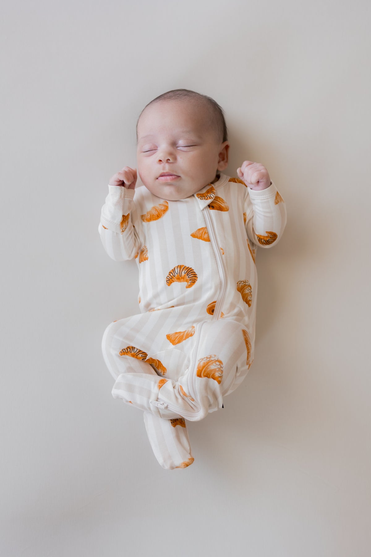 Baby sleeping wearing Zippered Footie with Croissant print on a beige background