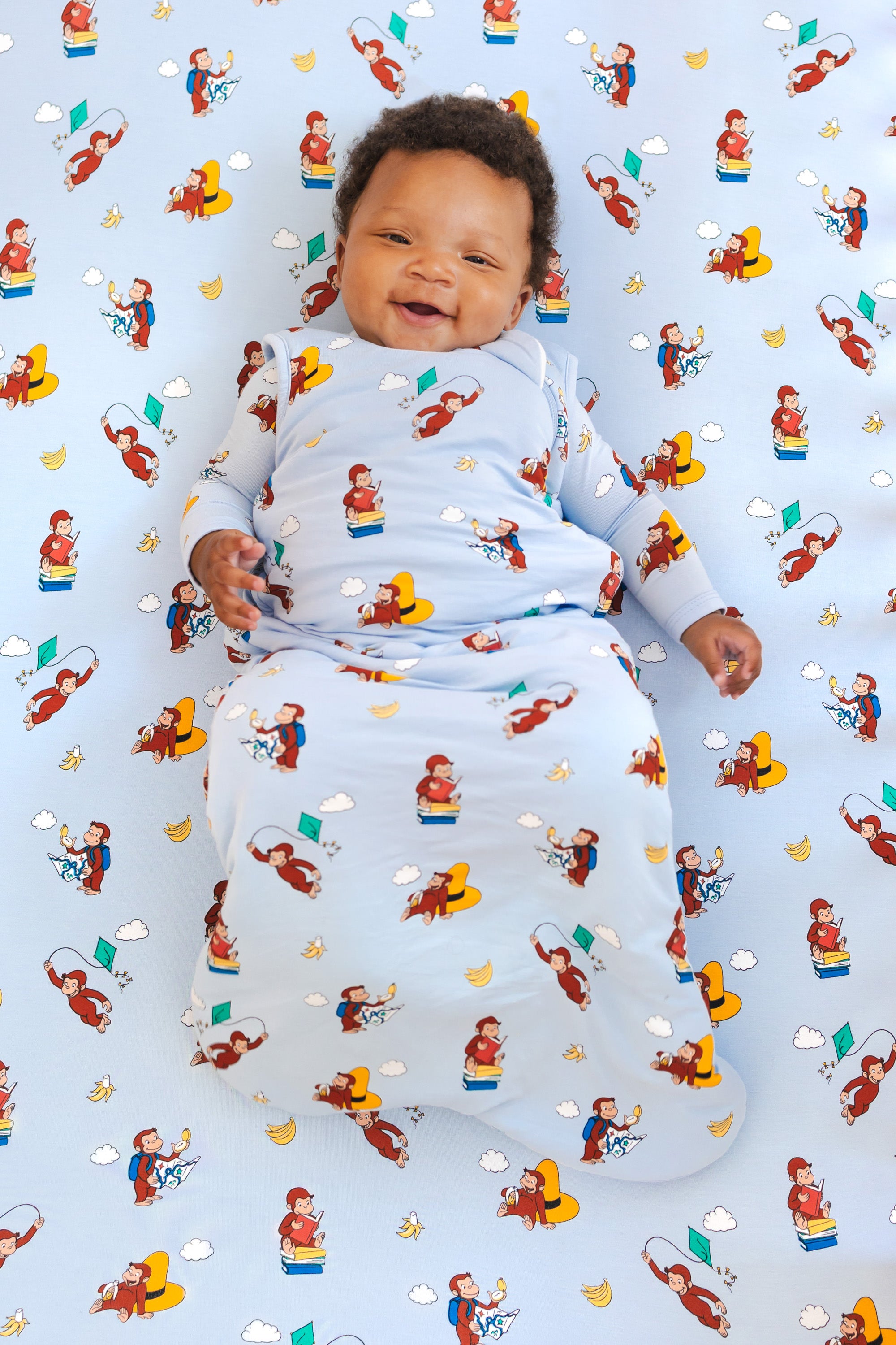 Baby smiling in Sleep Bag in Curious George 1.0 print lying on a similar print crib sheet