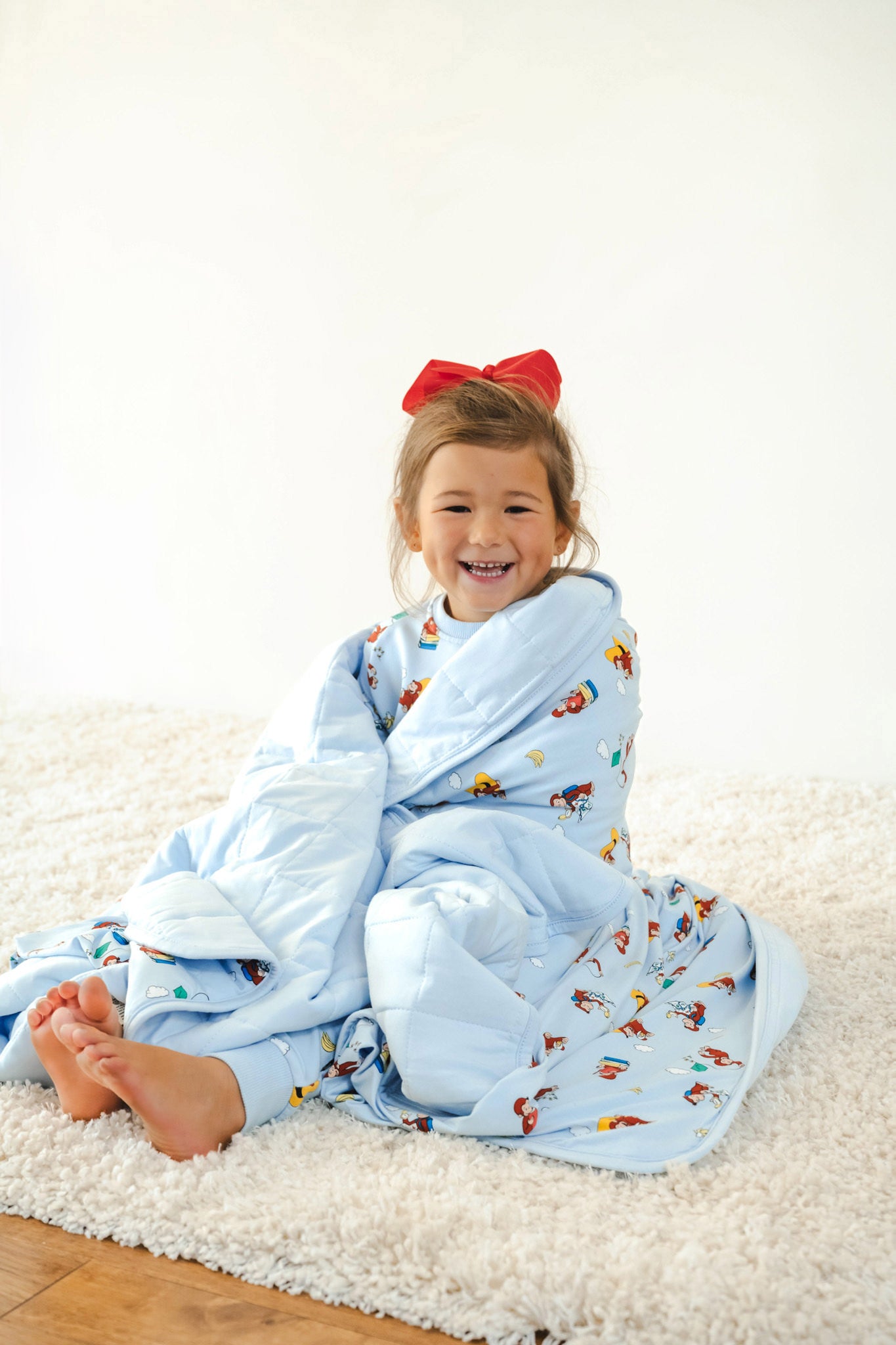 Girl smiling wrapped in Youth Blanket in Curious George 2.5 print wearing similar print long sleeve jogger set and red bow on head sitting on a white carpet
