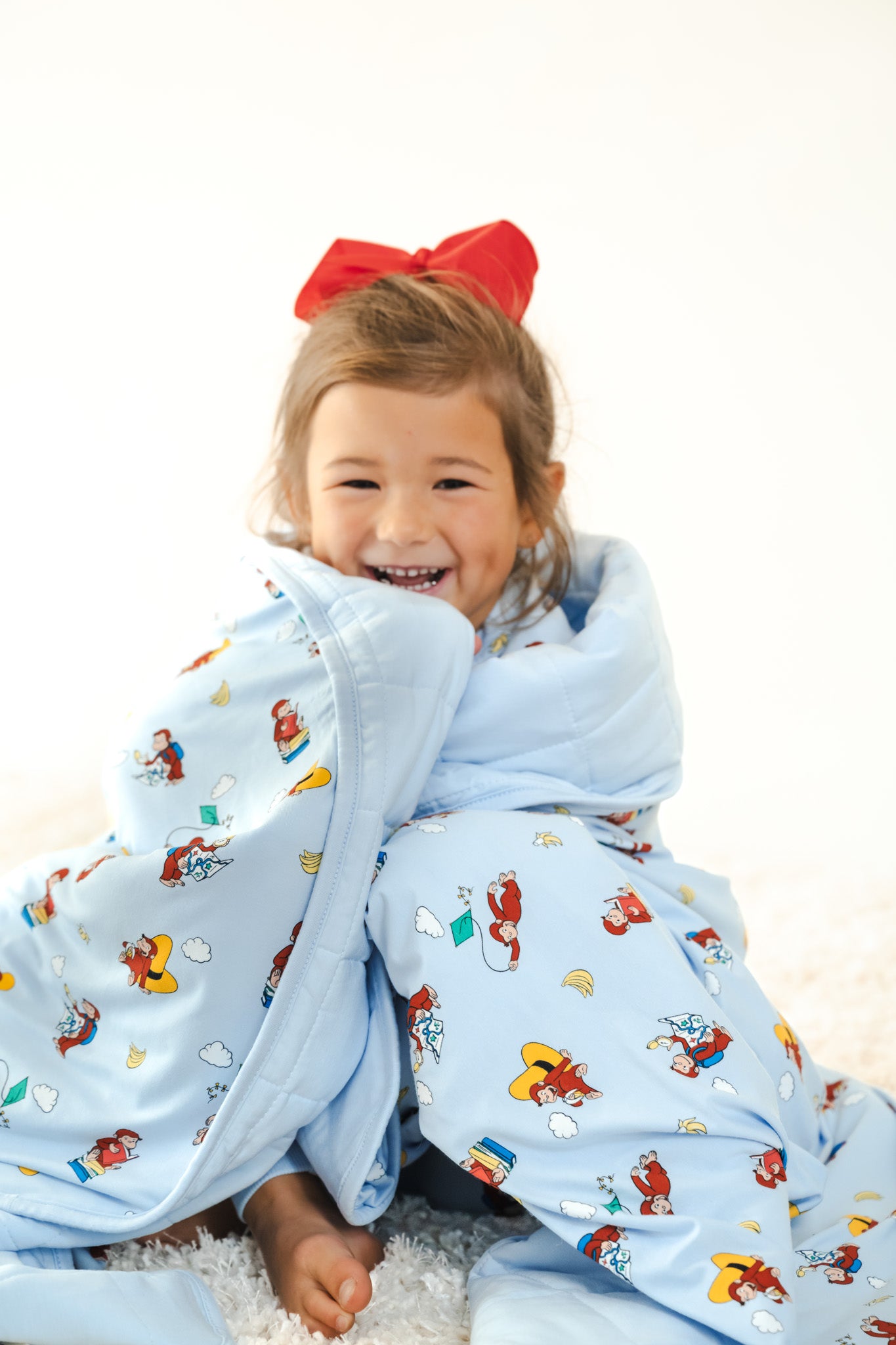 Girl wrapped in Youth Blanket in Curious George 2.5 print wearing red bow on head sitting on a white carpet