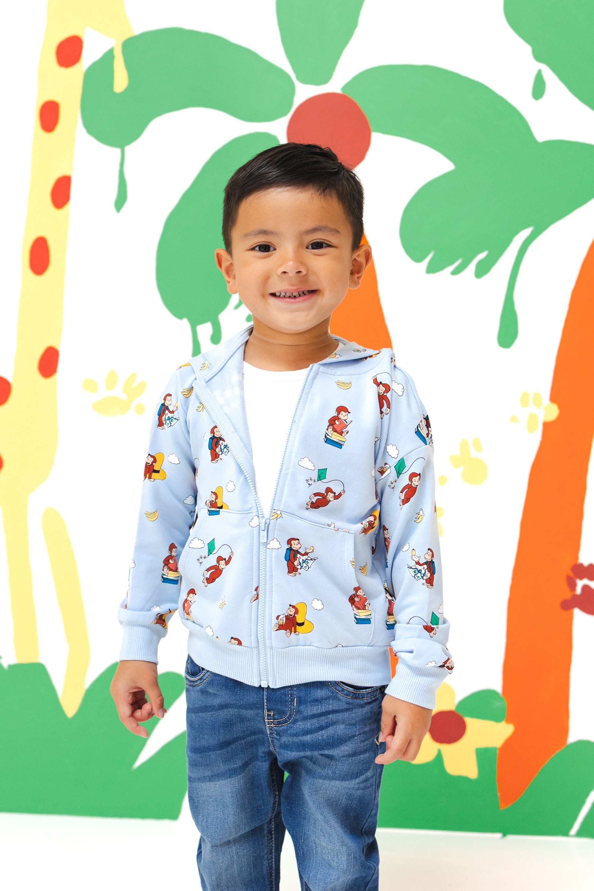 Boy smiling in Zip-Up Hoodie in Curious George print over a blue jeans standing in front of a colorful background