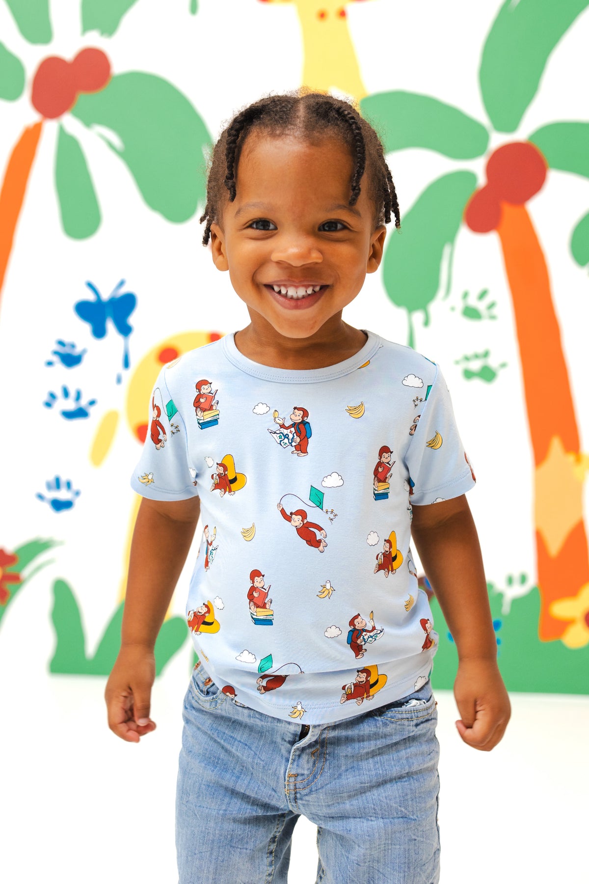 Girl wearing Toddler Basic Tee in Curious George print over a blue jeans smiling in front of a colorful background