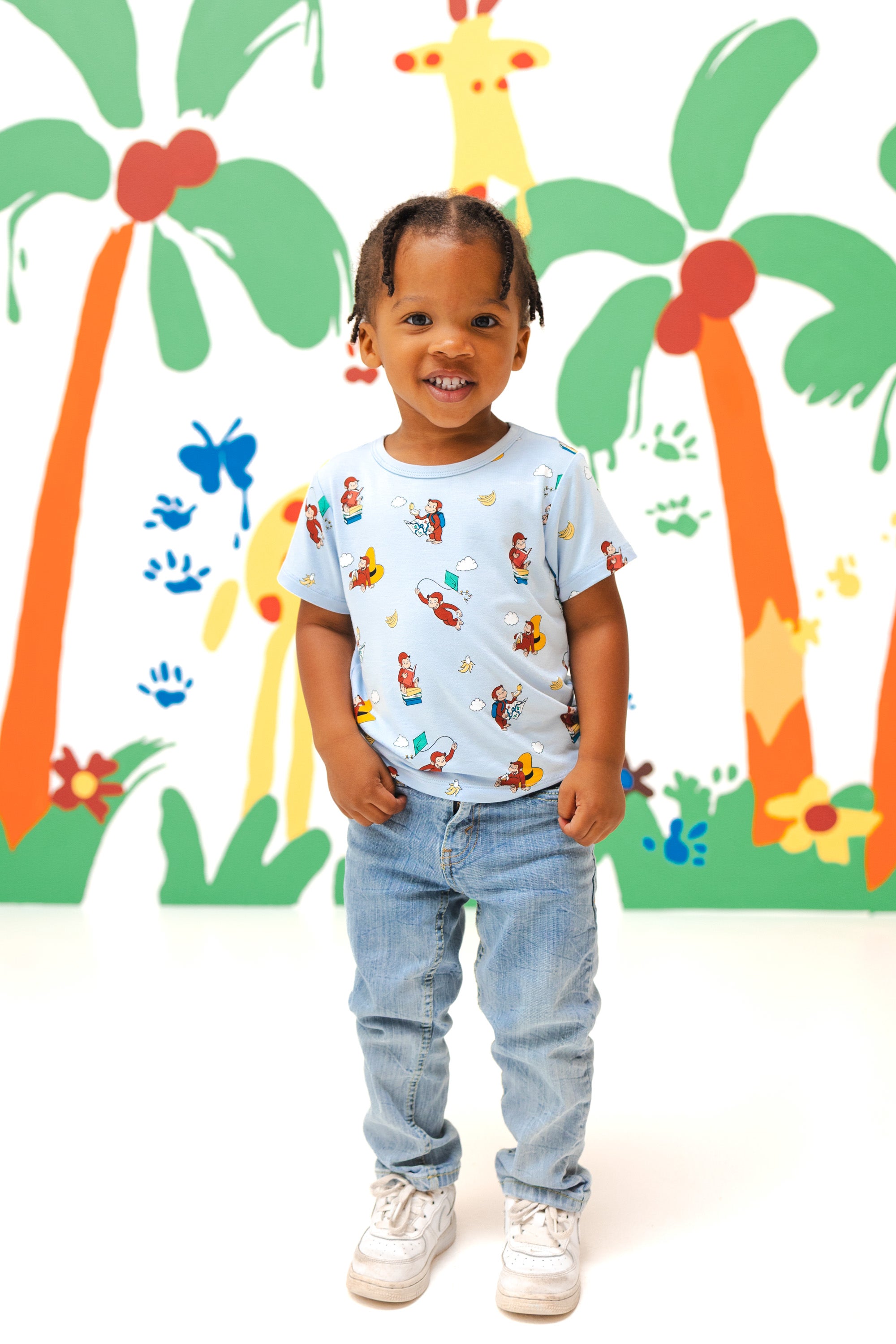 Girl posing in Toddler Basic Tee in Curious George print over a blue jeans smiling in front of a colorful background