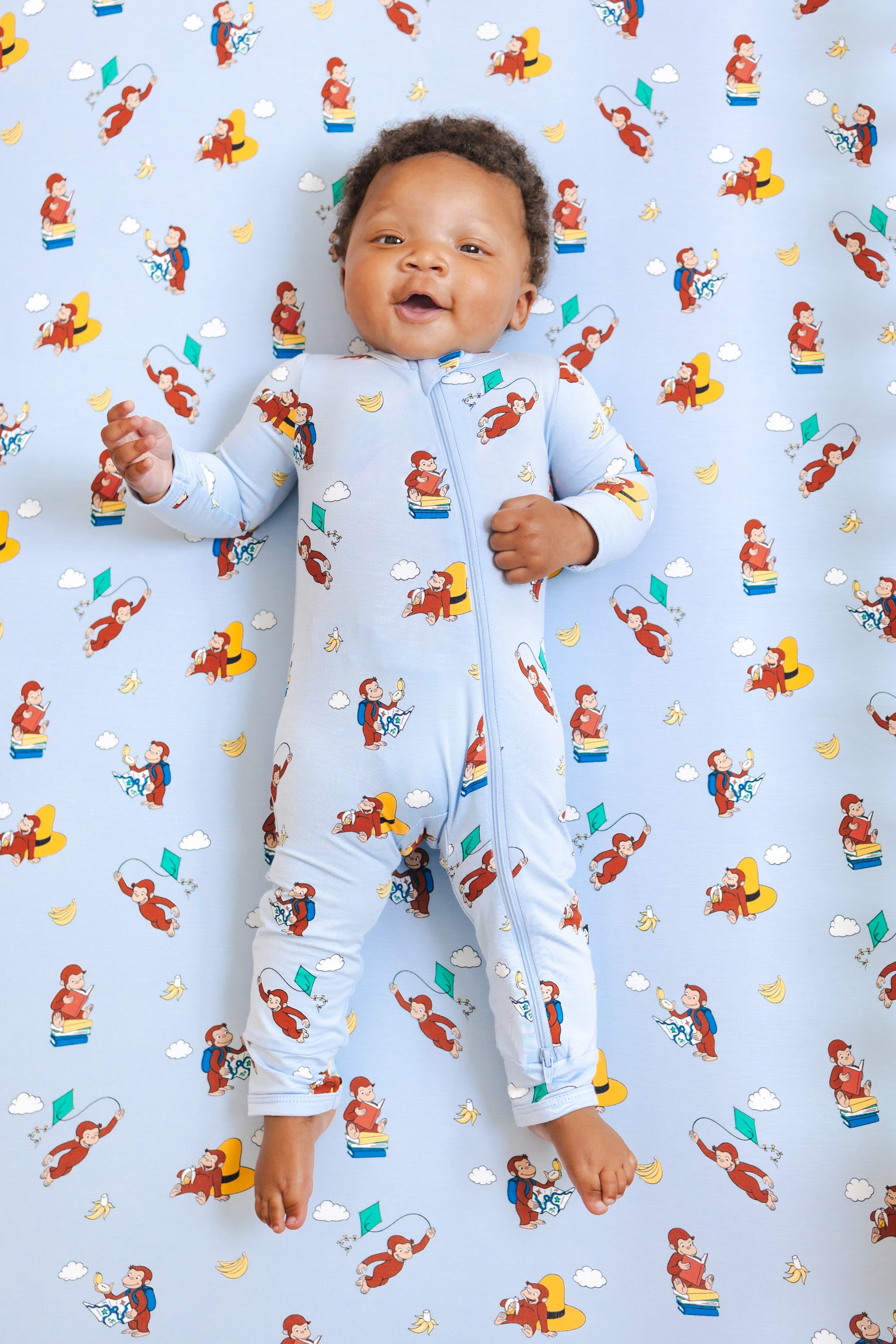 Baby wearing Zippered Romper in Curious George print lying over a similar print crib sheet