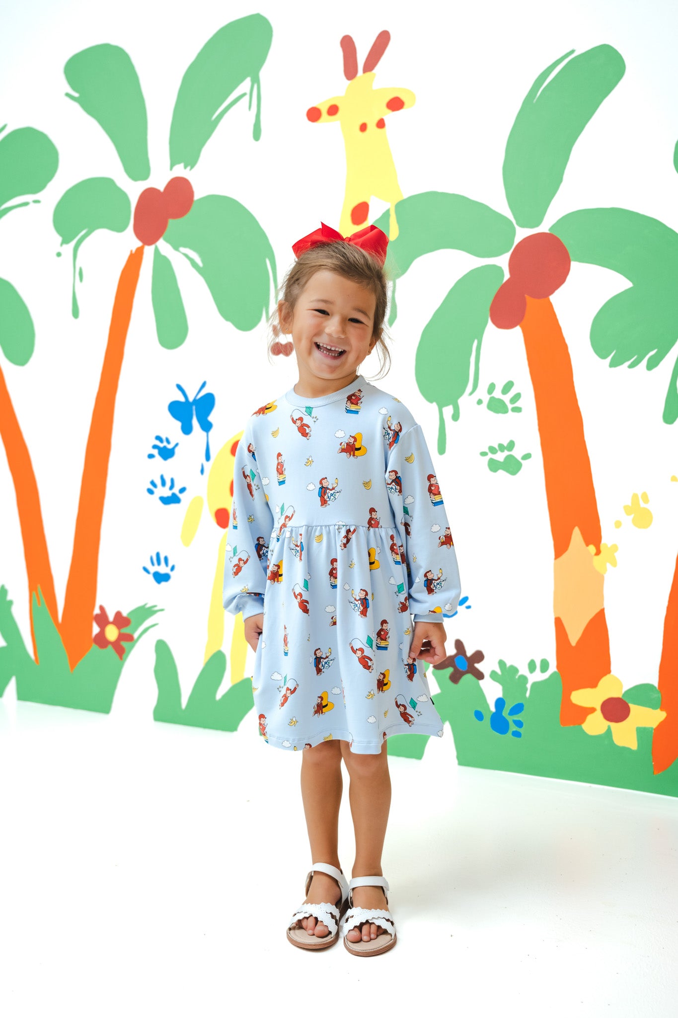 Girl wearing Long Sleeve Skater Dress in Curious George print with a red bow on head standing in front of a colorful background