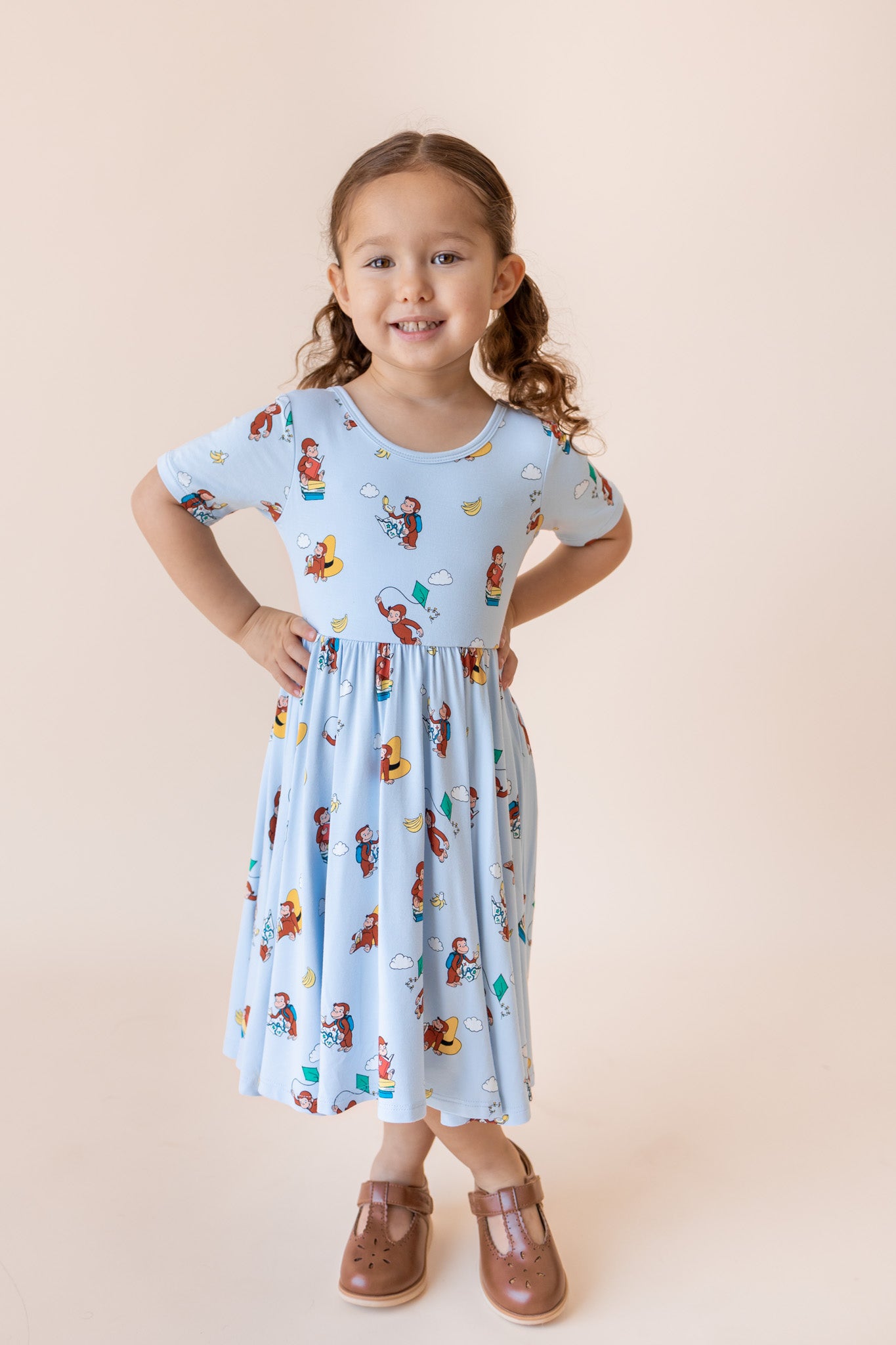 A young girl with curly brown hair in pigtails stands with hands on hips, wearing a light blue short-sleeve dress with a colorful cartoon print and brown mule shoes, against a solid pale peach studio background.