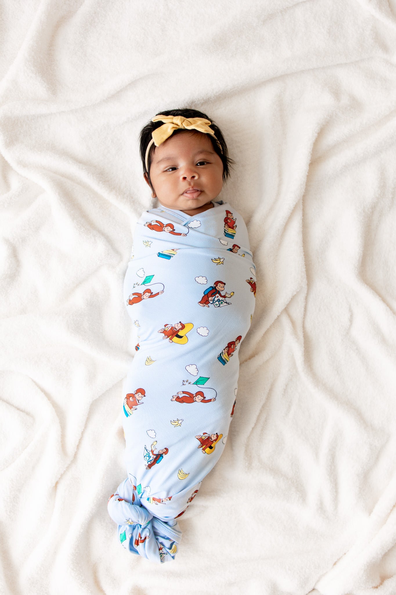 Baby swaddled in Blue Swaddle Blanket with Curious George print on a beige sheet