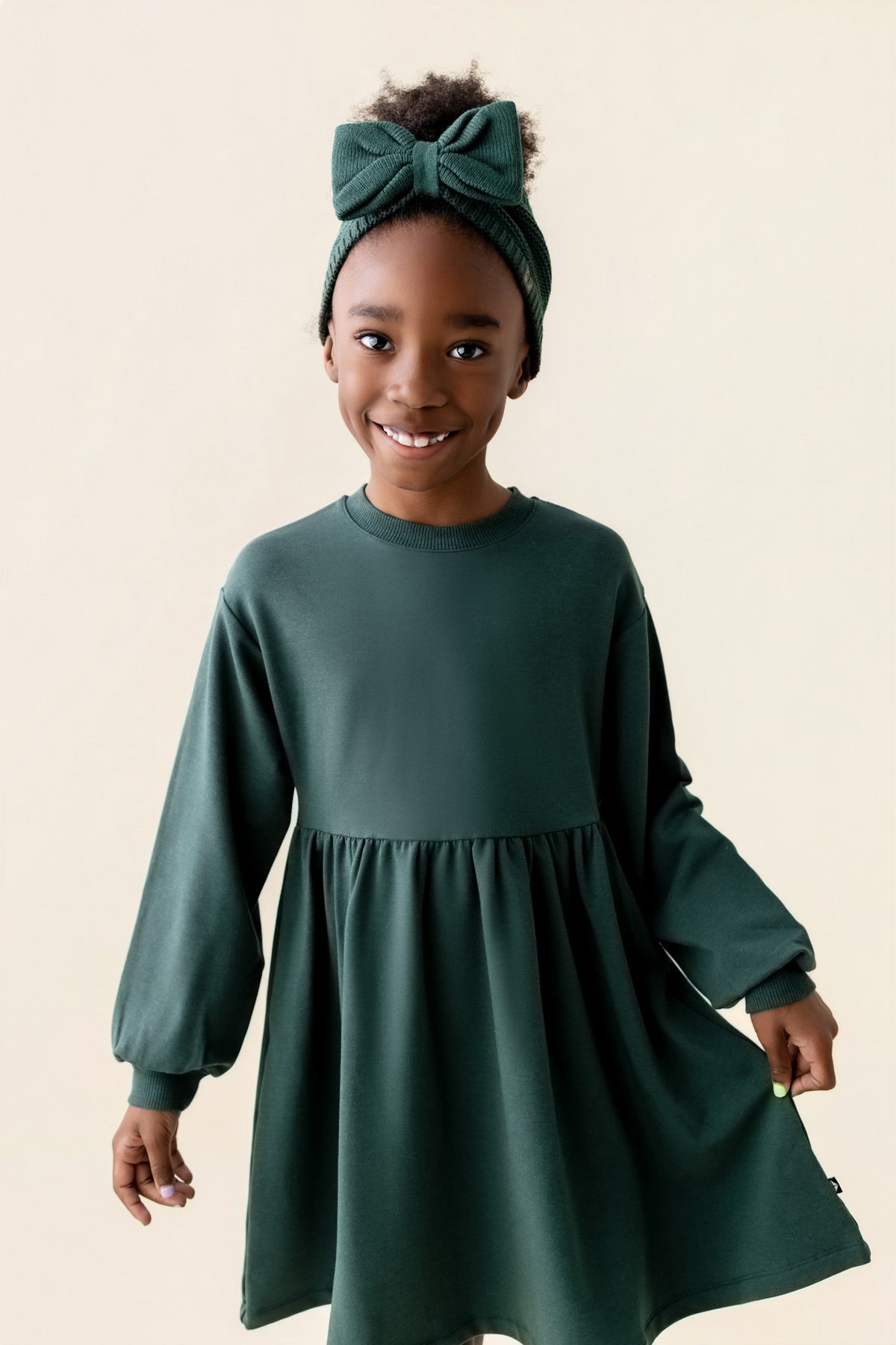 Girl wearing Long Sleeve Skater Dress in Cypress color with chunky knit knotted bow headband on a beige background