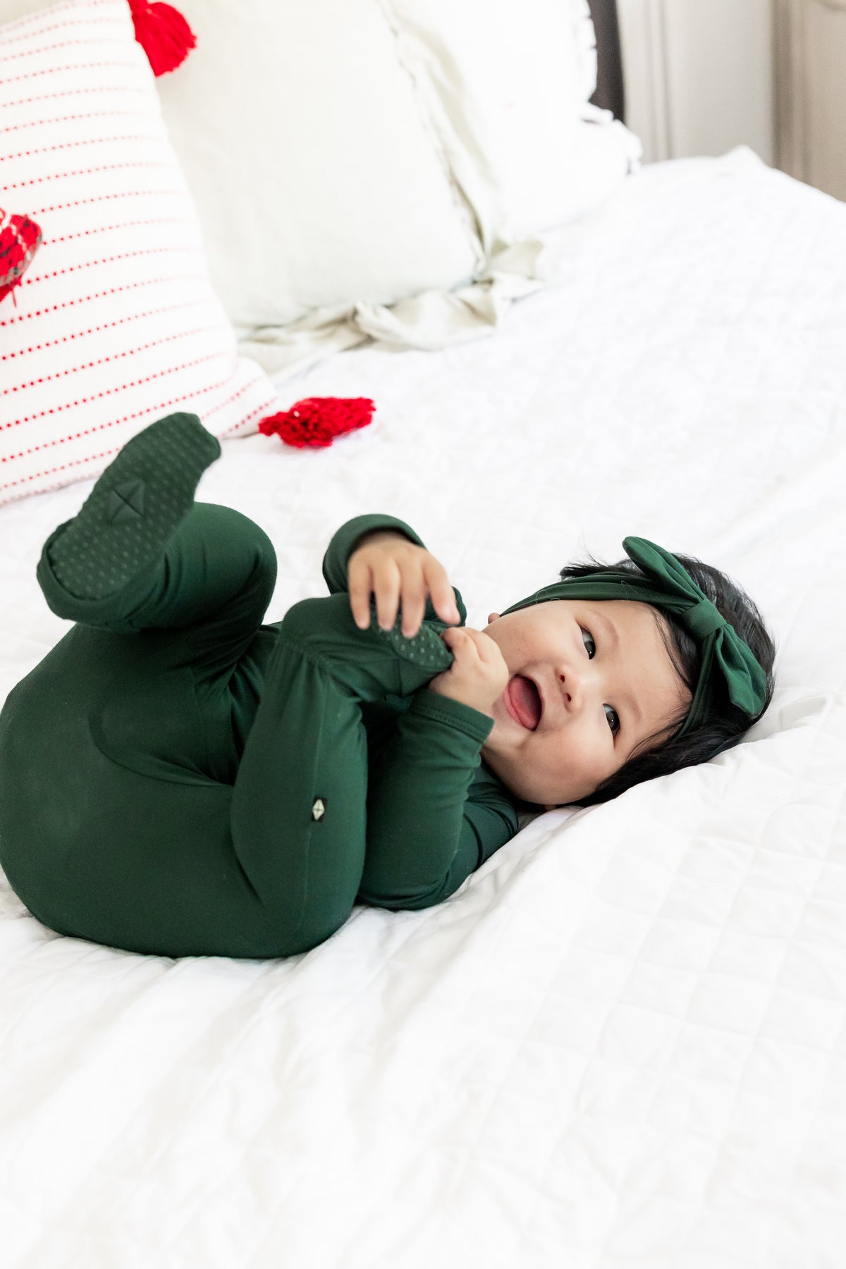 A baby lying on a white quilted bed wearing a dark green one-piece jumpsuit and matching green bow headband, with red decorative accents on the pillows.