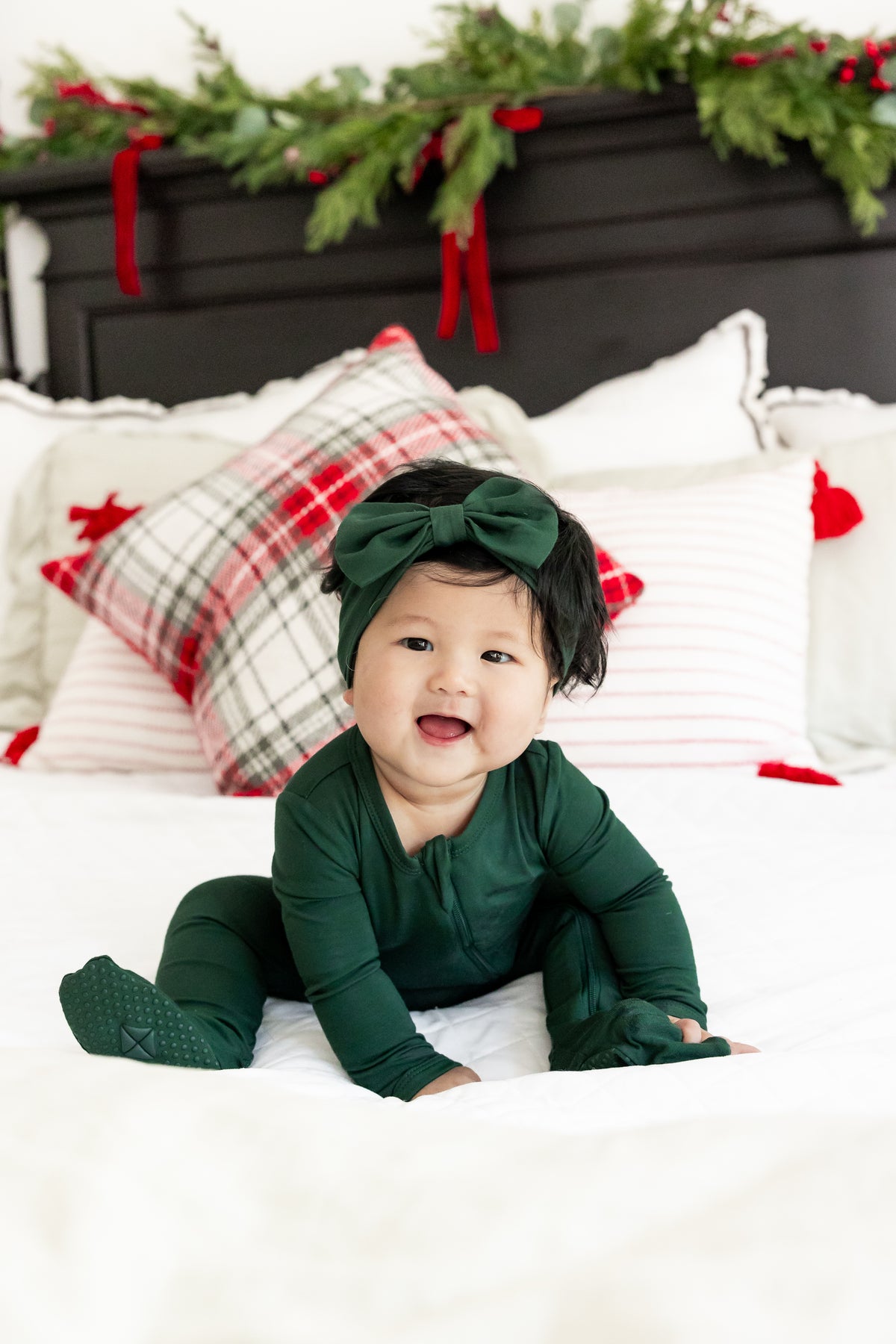 A baby wearing a dark green long-sleeve footed onesie and matching green bow headband sits on a white bed with holiday-themed pillows and a green garland in the background.